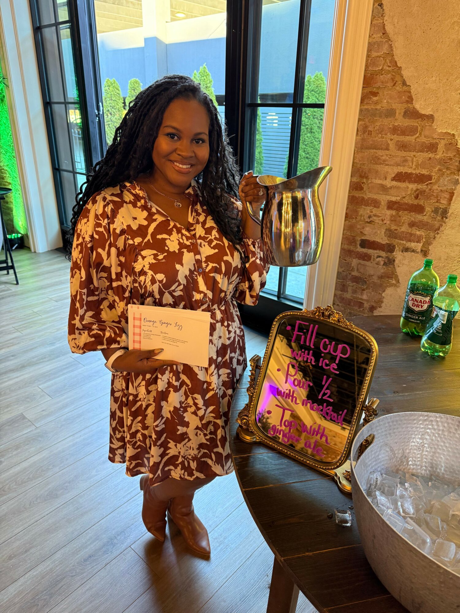 Woman in patterned dress holding a card, standing indoors near a table with drinks and a sign, window in background.