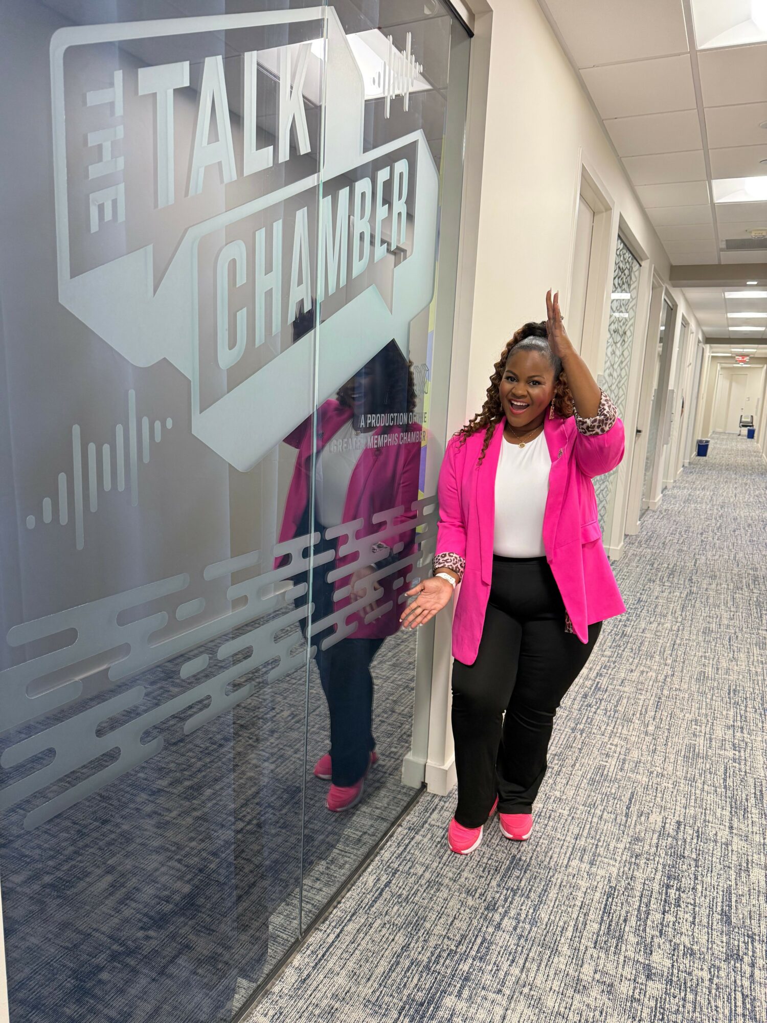 Woman in pink blazer and black pants standing next to a glass wall with text, waving and smiling.