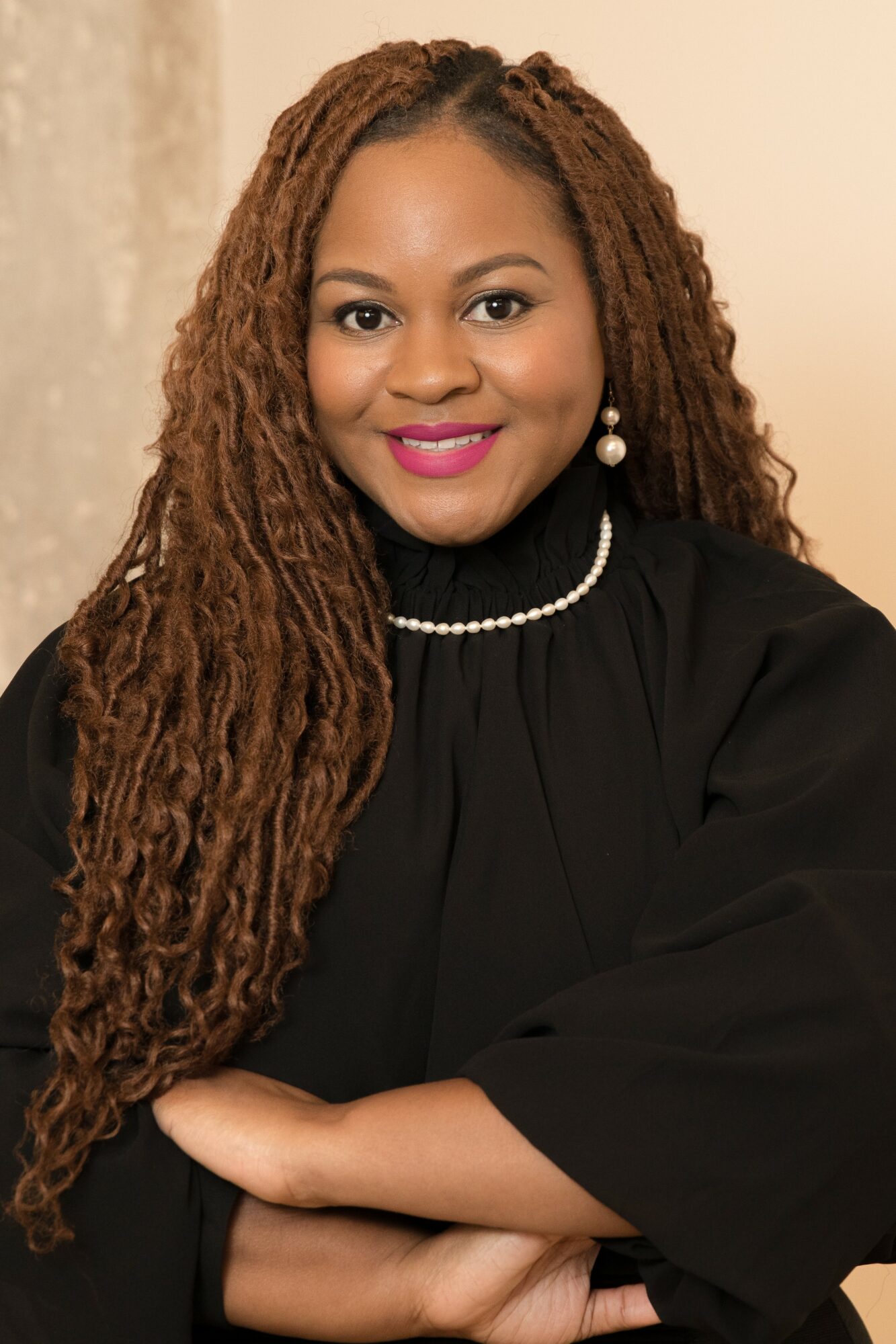 Portrait of a woman with long, curly hair, wearing a black top, pearl earrings, and a pearl necklace, smiling.