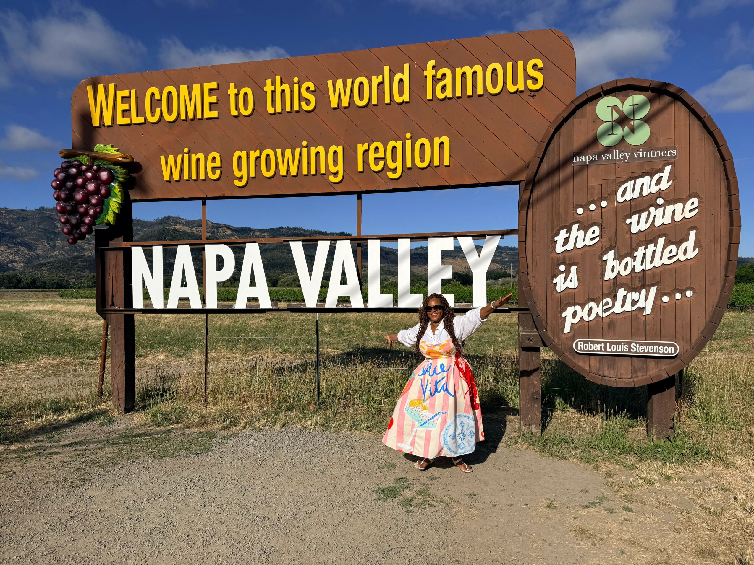 Person in colorful dress standing in front of signs for Napa Valley wine region, with blue sky and clouds.