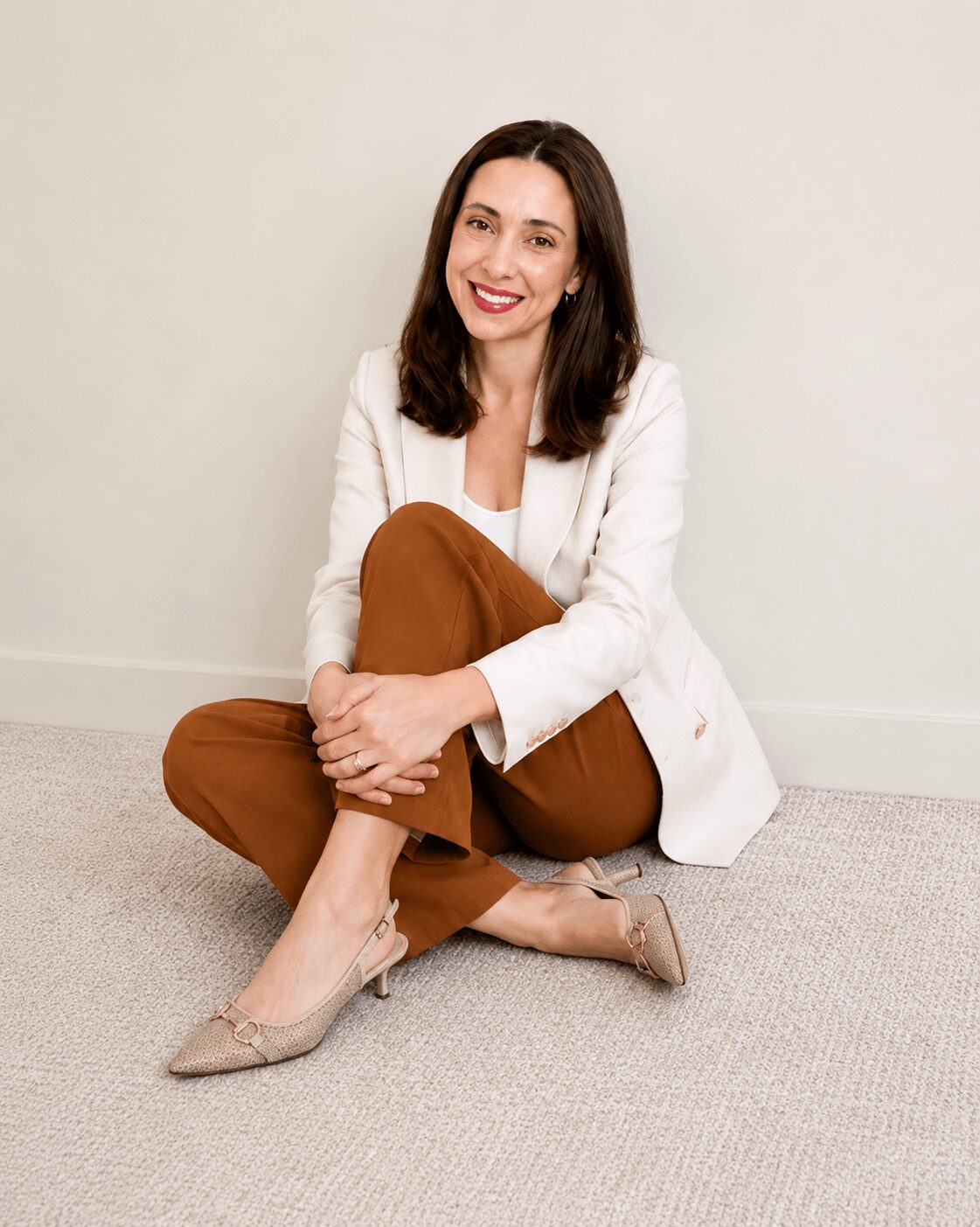 Woman sitting on floor with crossed legs, smiling, wearing white blazer, brown pants, and beige heels, against plain wall.