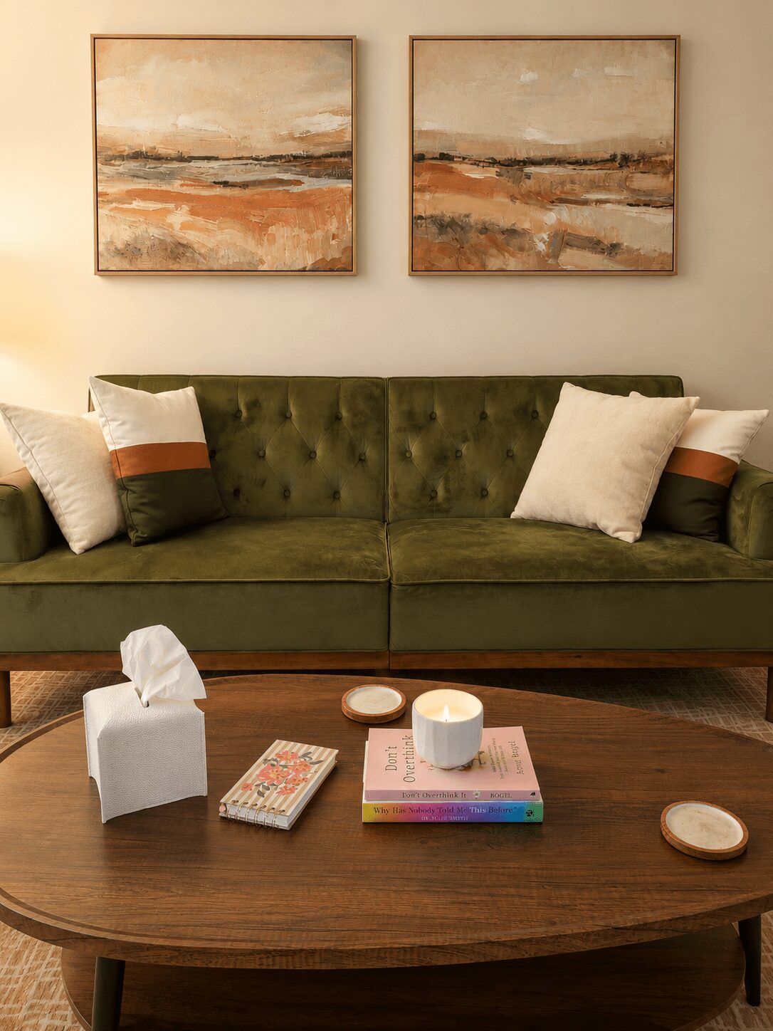 Living room with a green sofa, two white pillows, and abstract landscape paintings above. Wooden coffee table with tissue box, books, and cups.