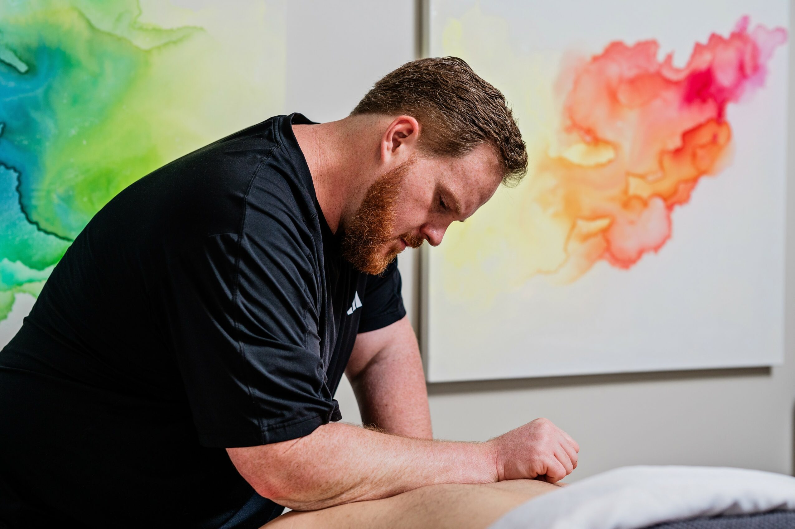 Man with beard leaning on a table, looking down, with colorful abstract paintings in the background.
