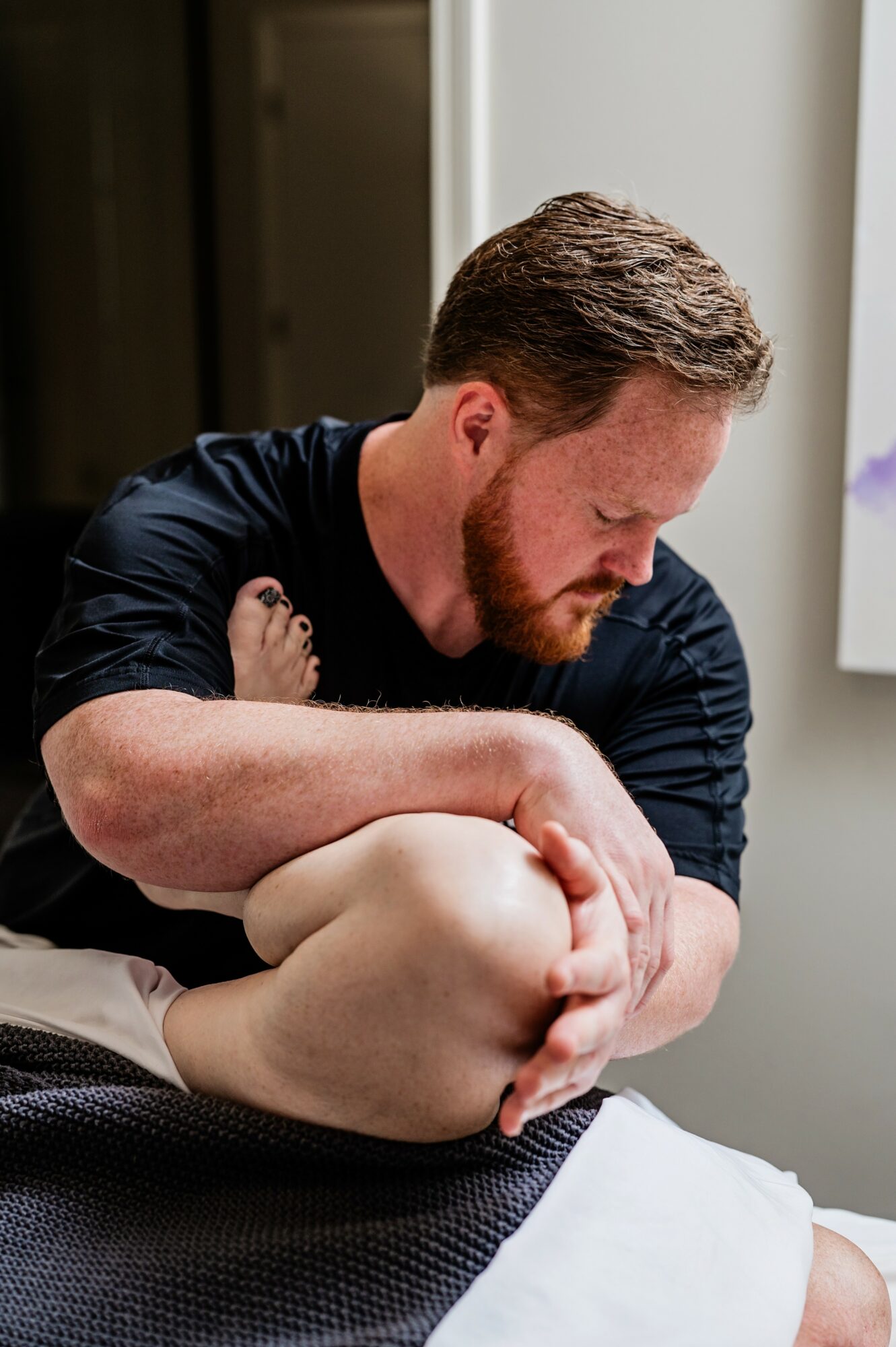 Man with red hair and beard holding a person's knee, sitting indoors near a window, focused on the knee.