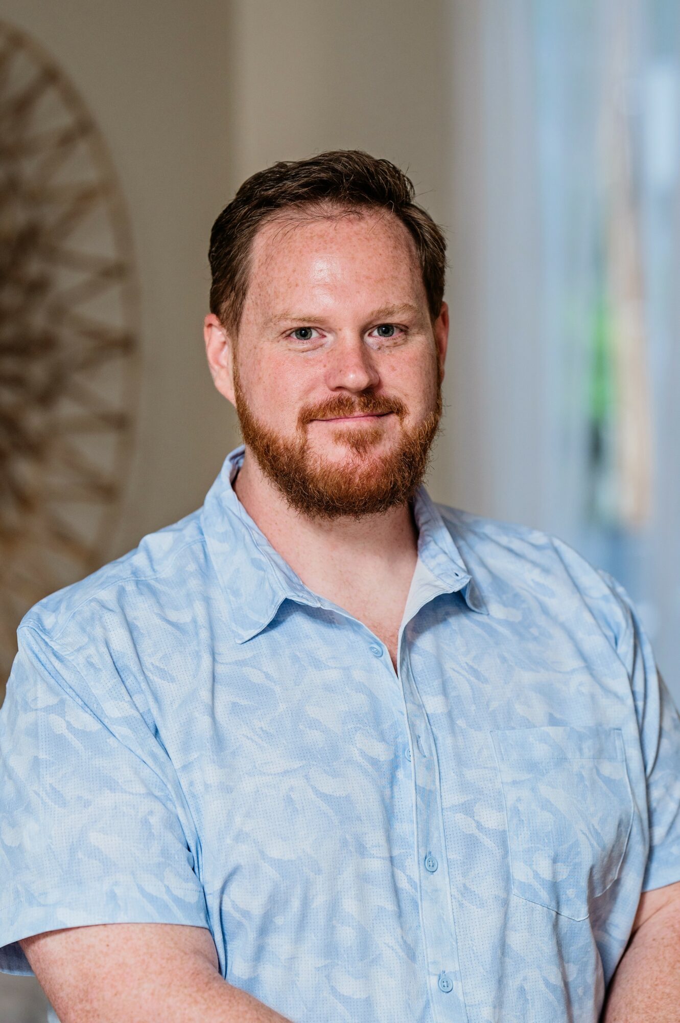 Man with short brown hair and beard wearing a light blue shirt, smiling indoors with a blurred background.