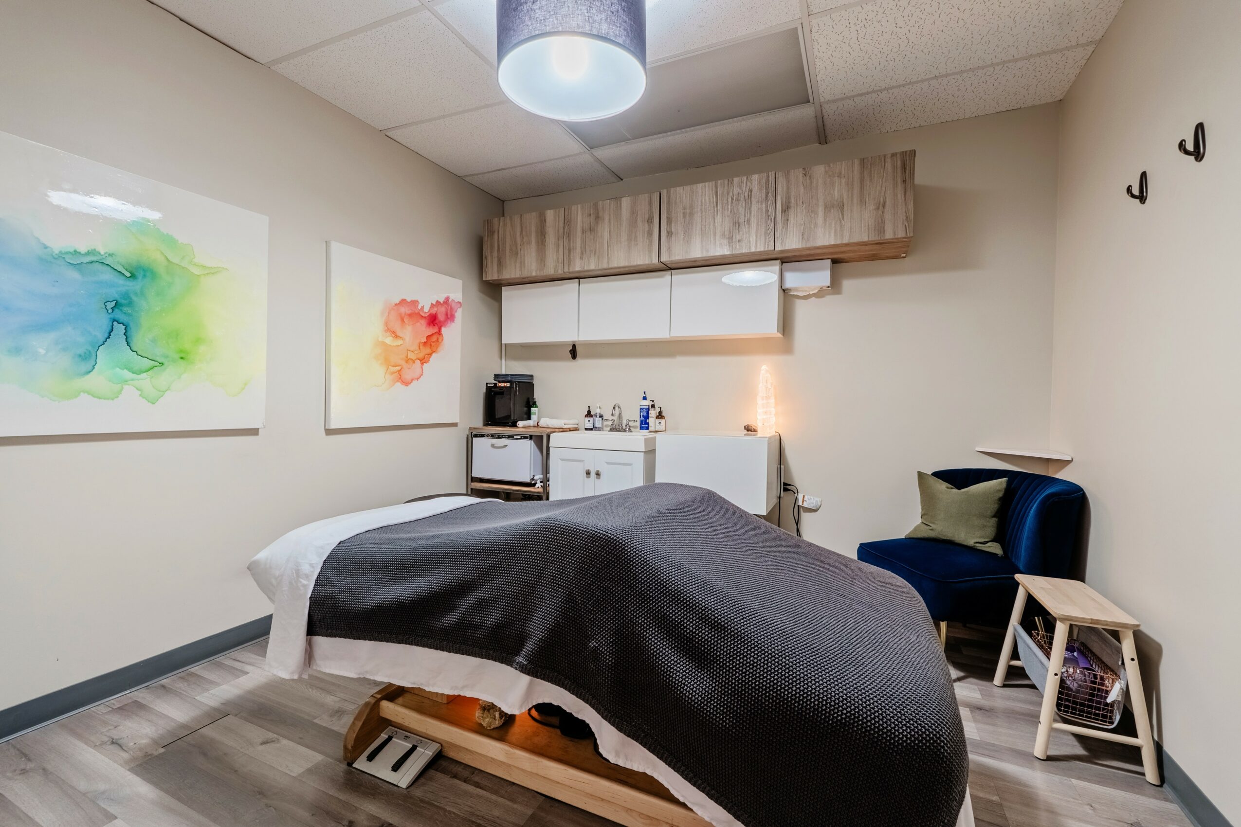 Hospital room with bed, artwork on walls, and a chair with a small table nearby.