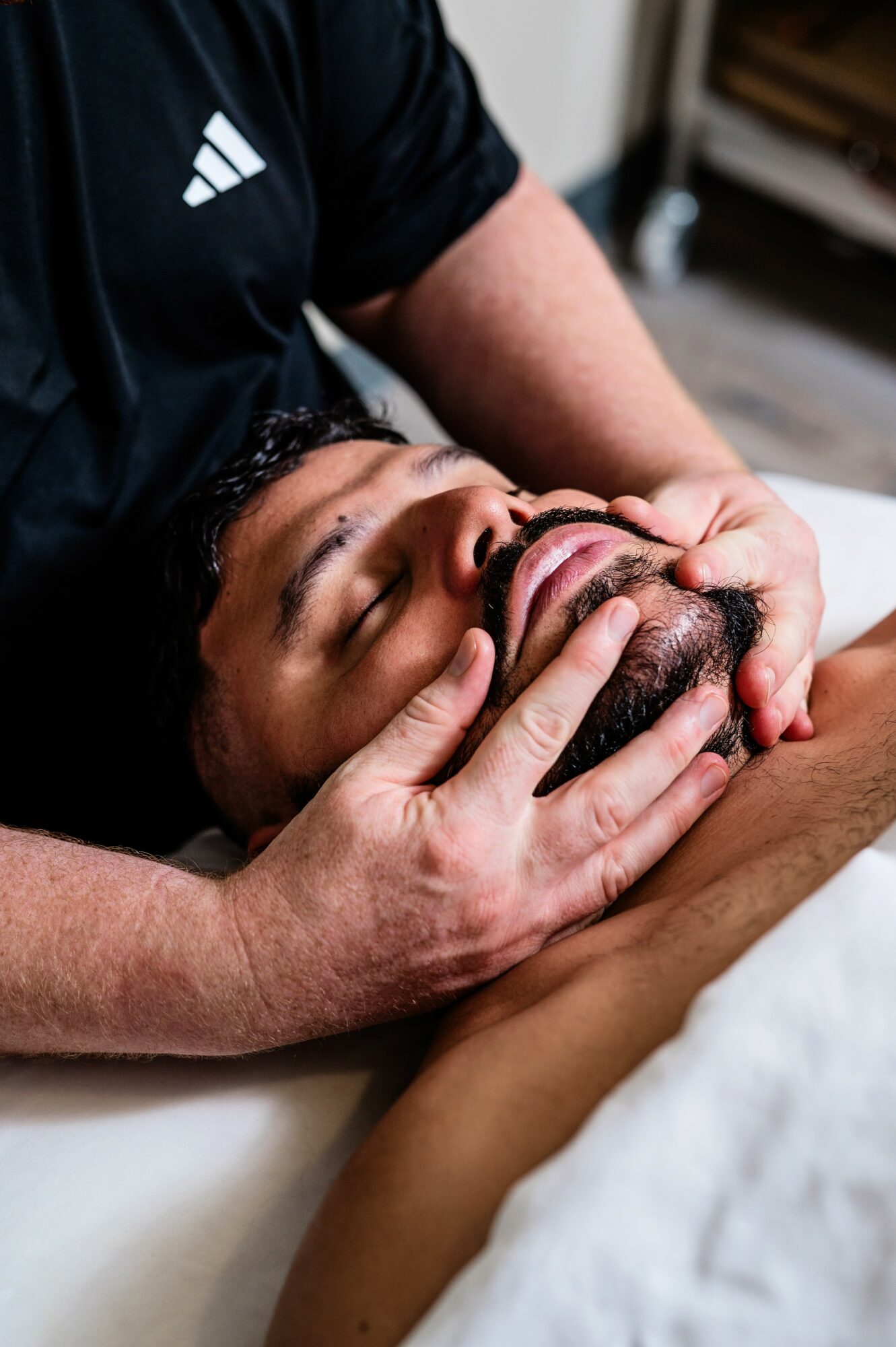 Person receiving a facial massage with hands on face, lying down with eyes closed, in a clinical setting.