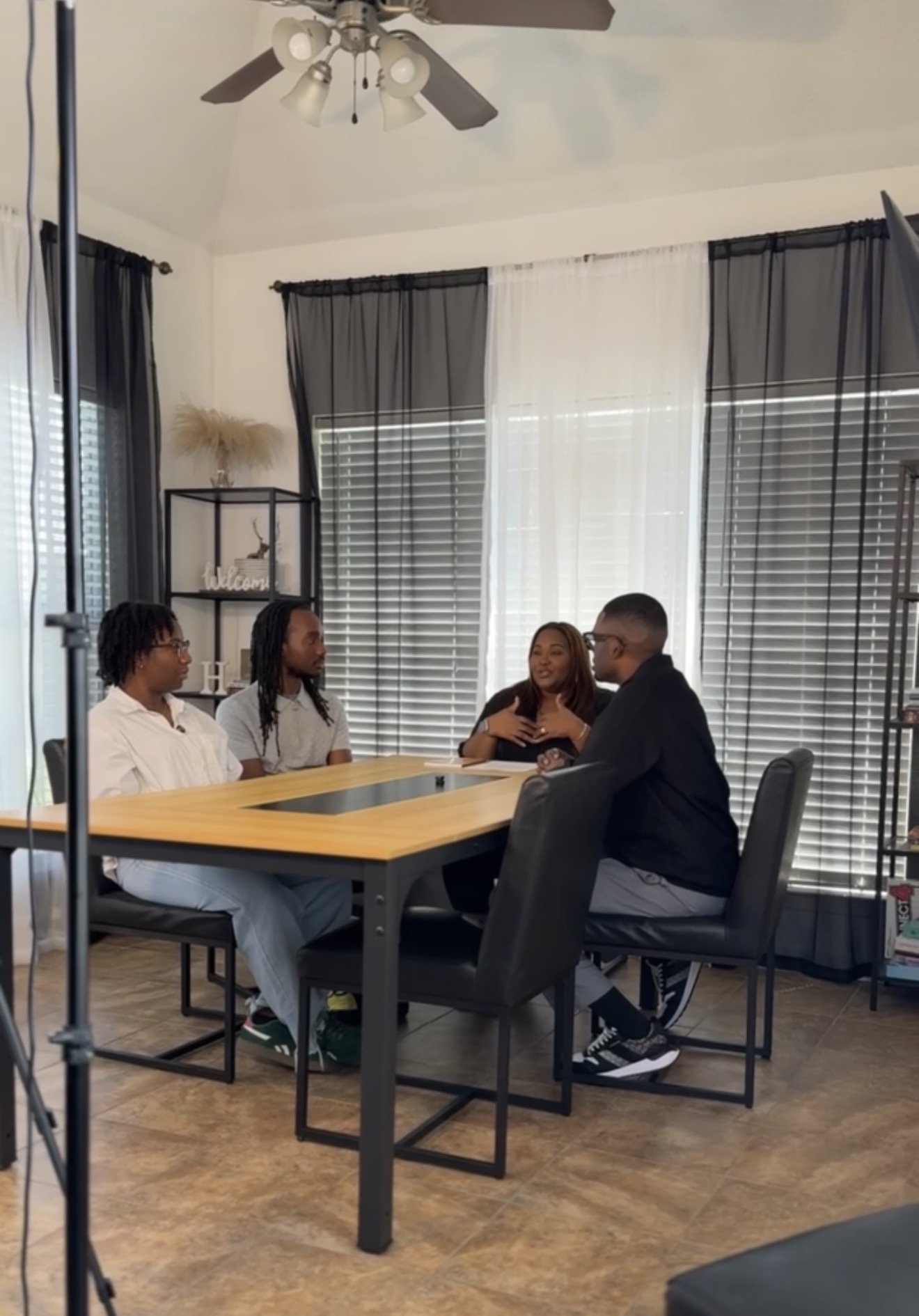 Four people sit around a table in a room with large windows and curtains, engaged in conversation.