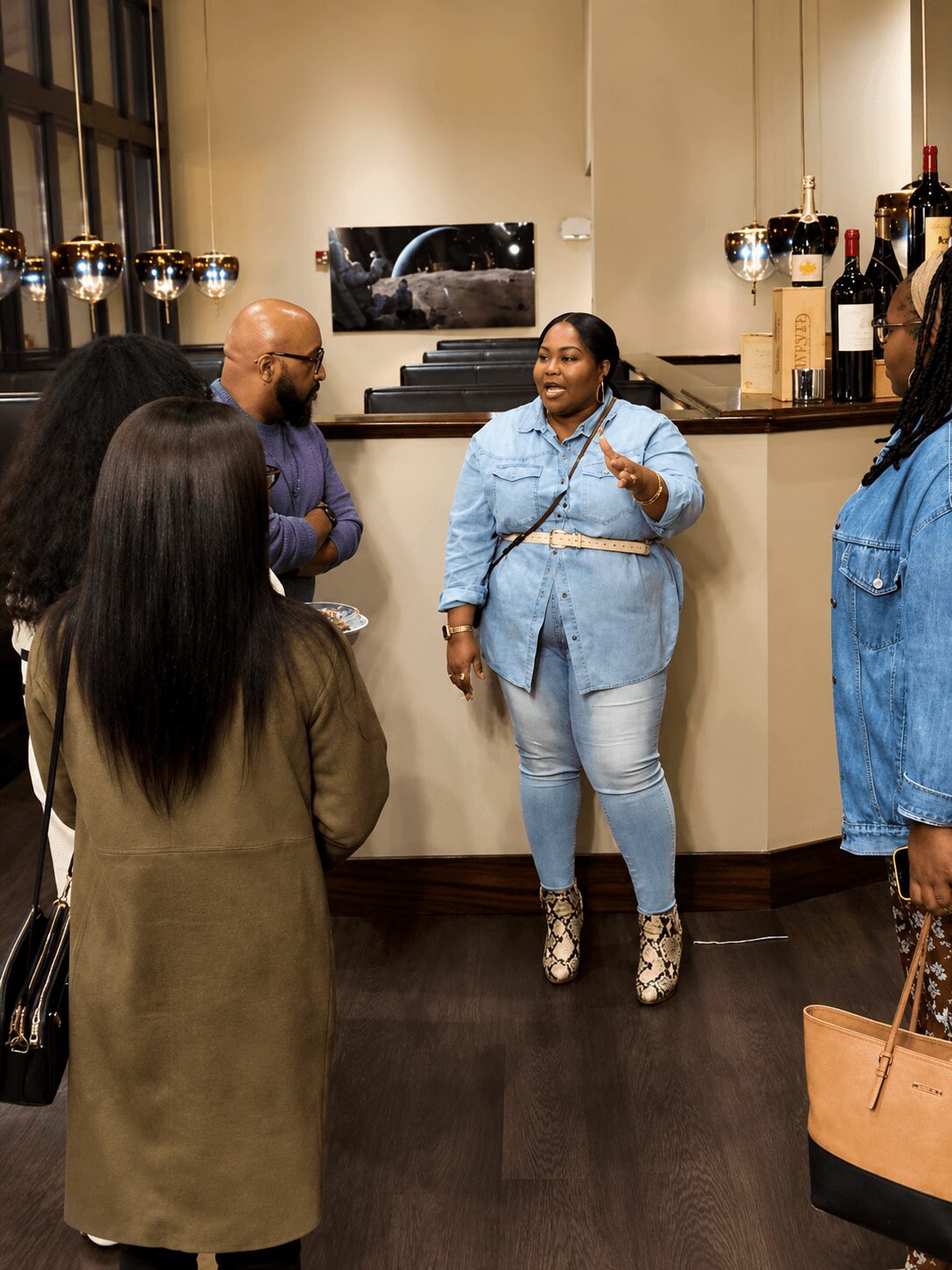 Group of people talking in a room with a bar counter, bottles, and glasses, with a woman in light blue jeans and jacket speaking.