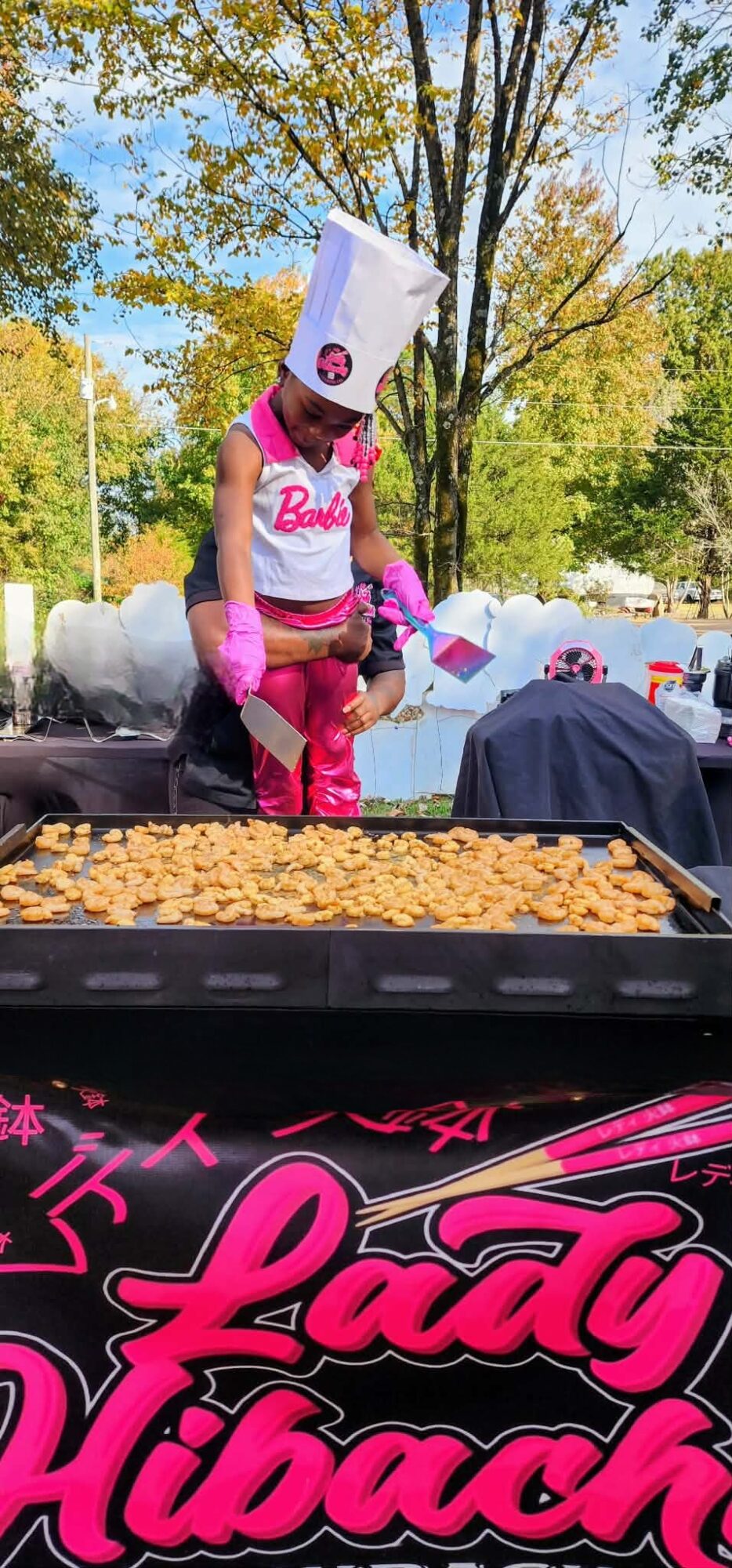 Person wearing a box on their head, pink outfit, standing outdoors near a tree and a table with food.