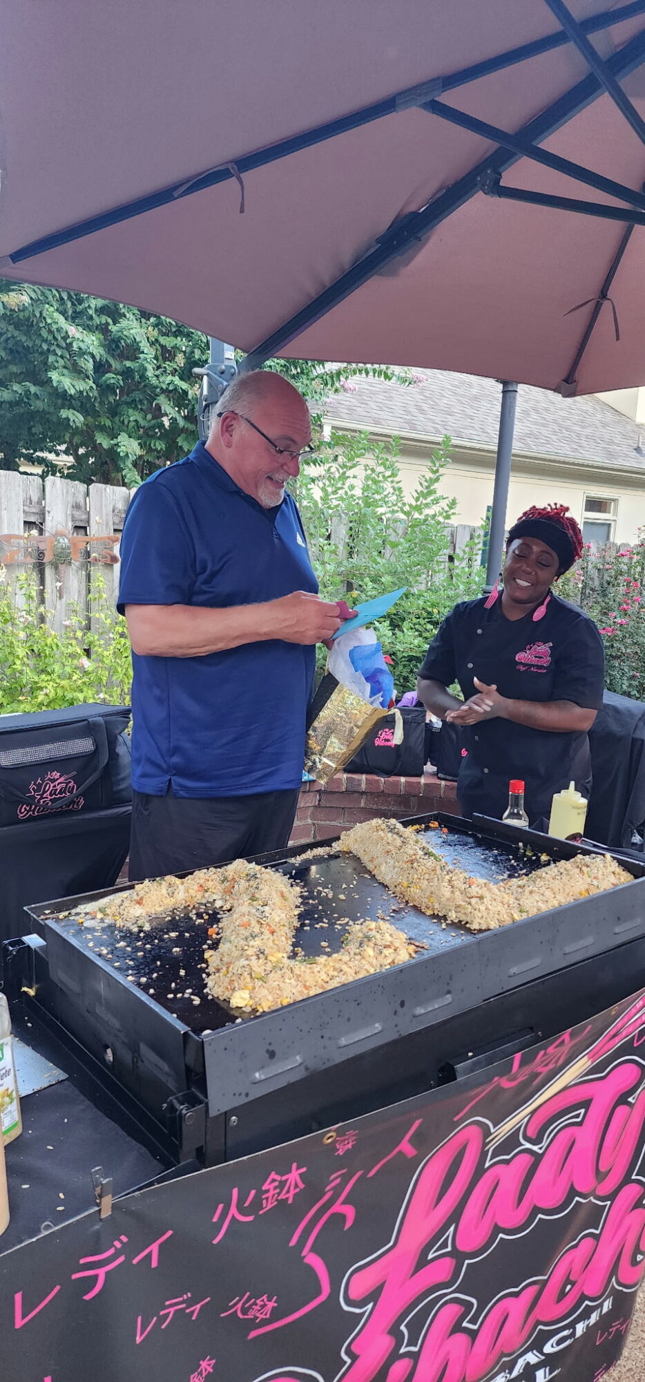 Two people cooking food outdoors under a large umbrella, with trays of food on a grill, surrounded by greenery.