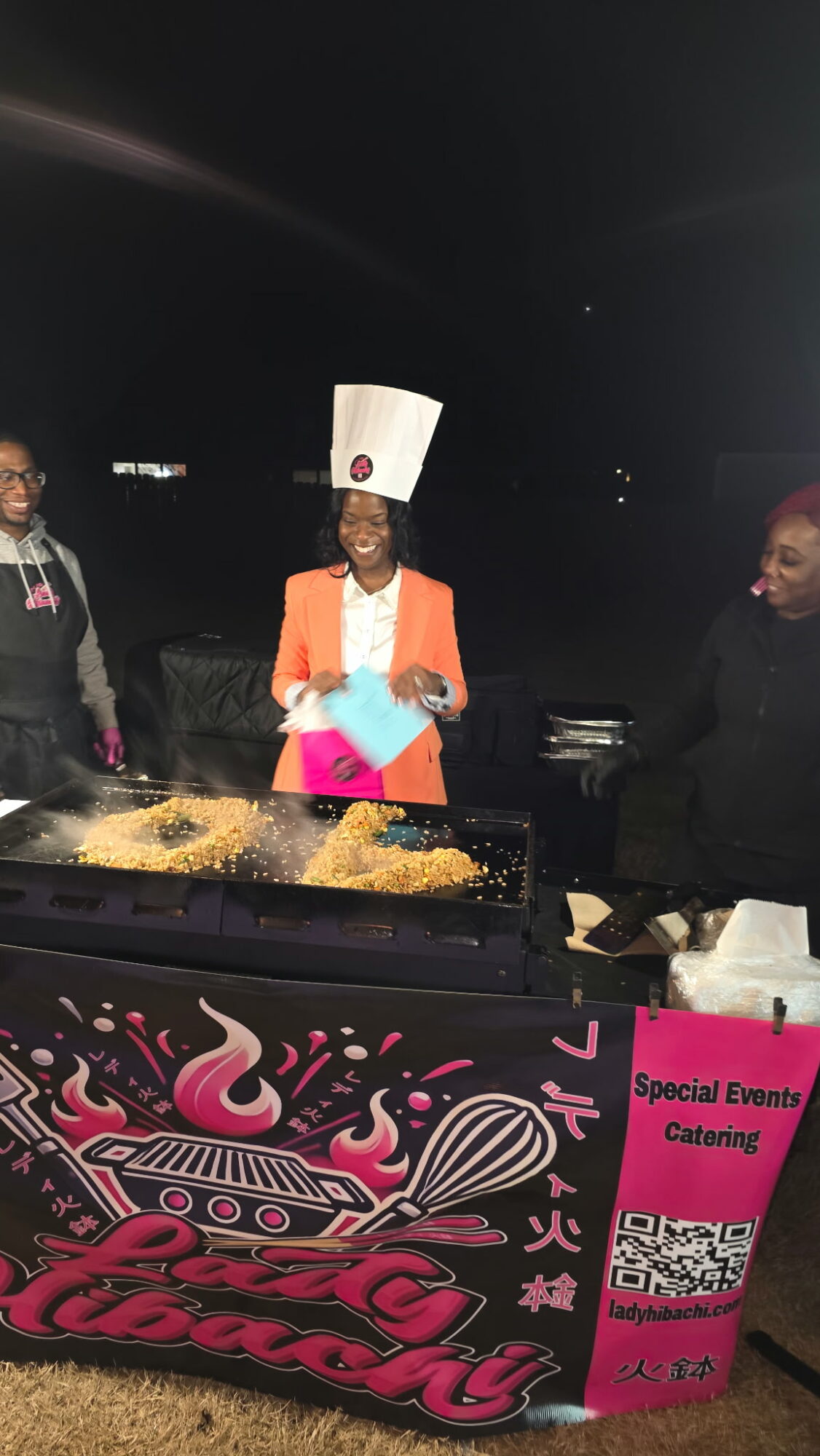 Three people standing outdoors at night, one wearing a tall white hat and orange blazer, with a pink and blue object, in front of a decorated table.
