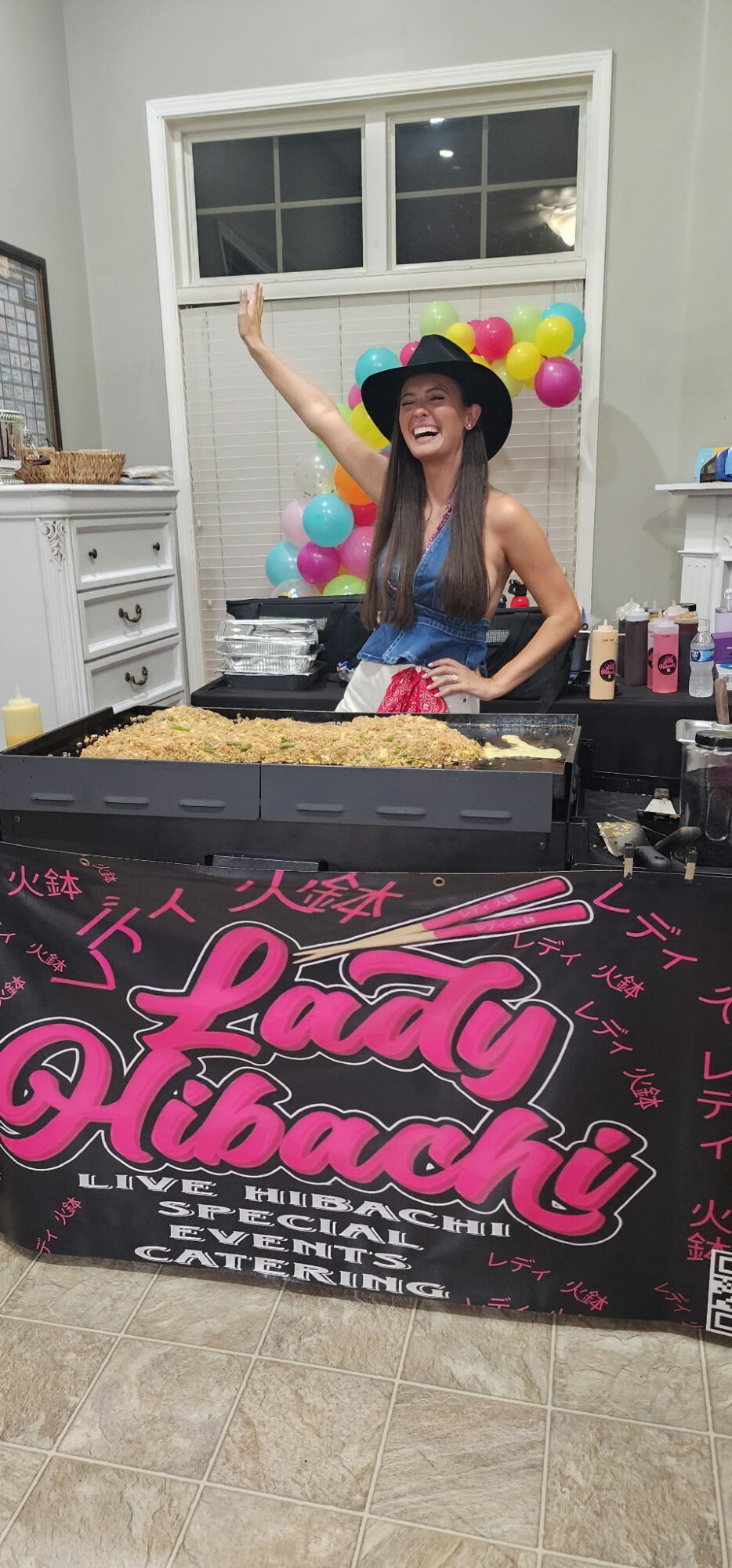 Woman wearing a hat and glasses standing behind a table with colorful balloons, a sign reading 'Lady Diaboli,' and food items.