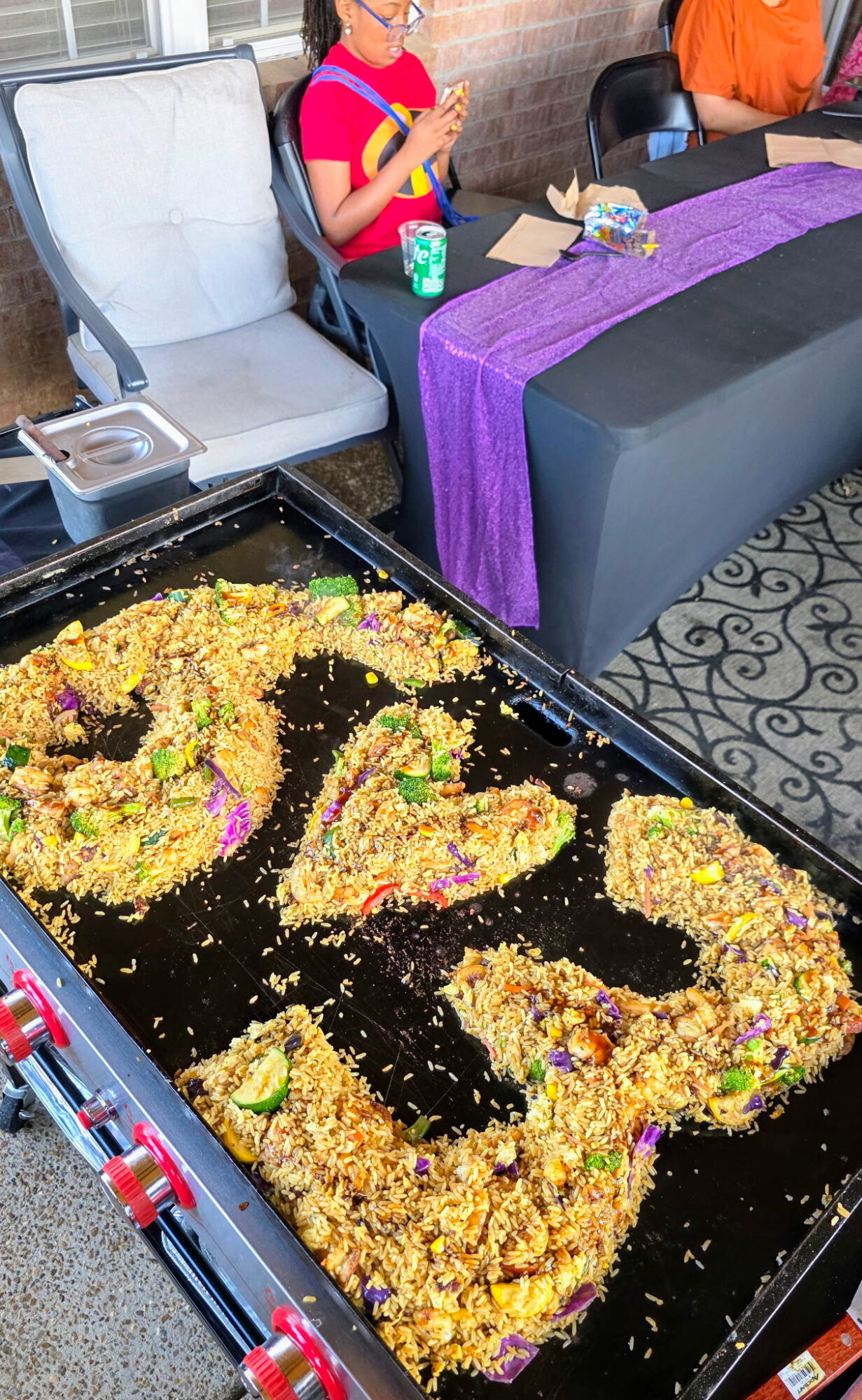 Tray of colorful rice and food shaped into the word 'LOVE' on a table with a purple tablecloth, people in background.