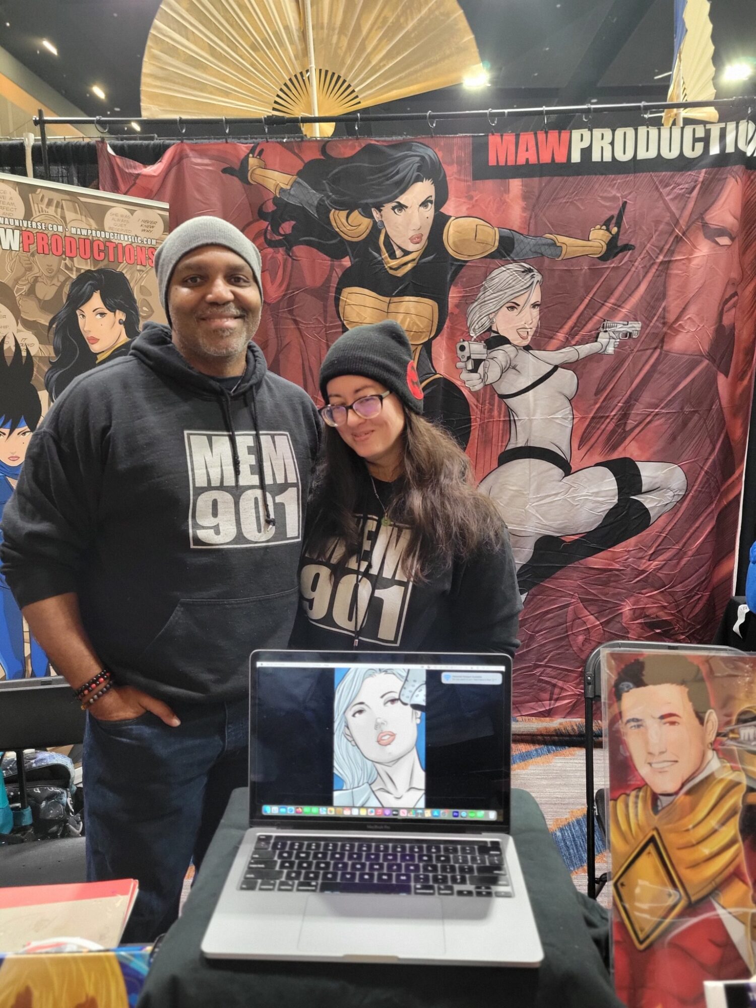 Two people standing behind a table with a laptop and artwork, smiling at the camera, at a booth with animated character backdrop.