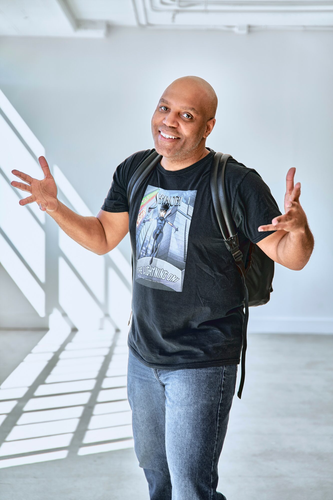 Smiling person with a backpack gesturing with hands, standing in a bright, modern space with shadows on the floor.