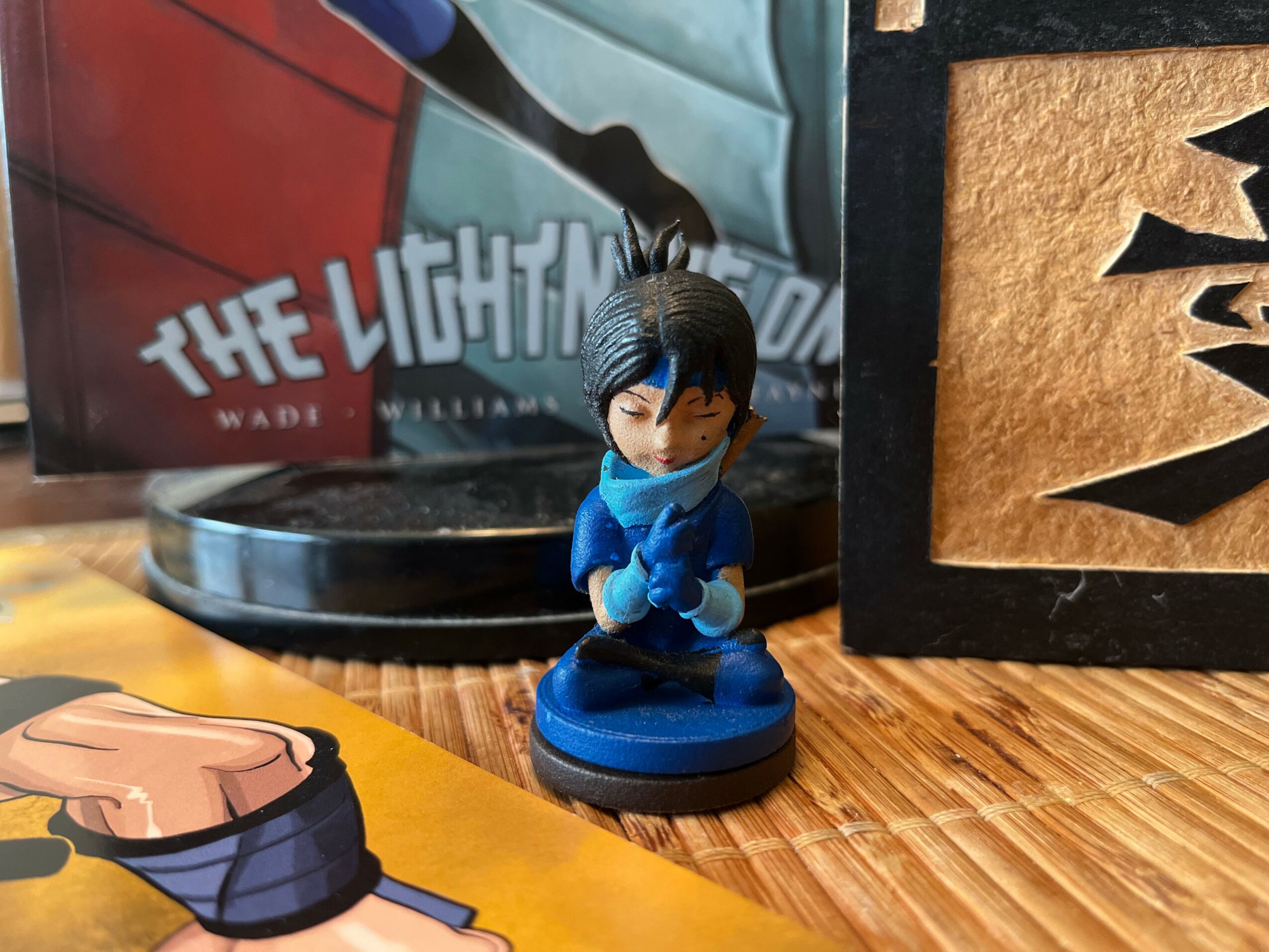 Small figurine of a character with black hair and blue clothing, standing on a round blue base, on a wooden surface.