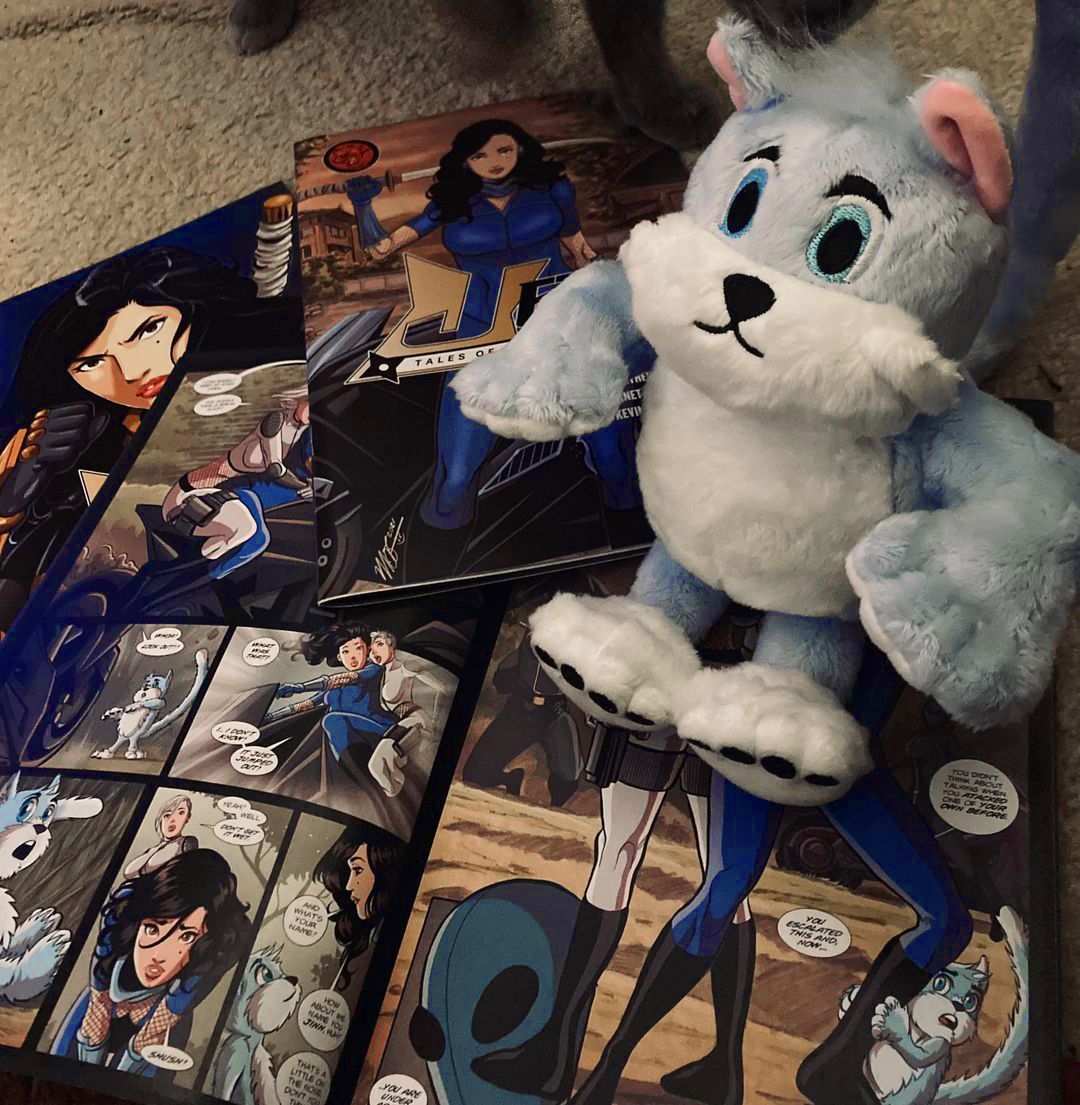 Plush toy of a white and gray cat with blue eyes, surrounded by comic books on a table.