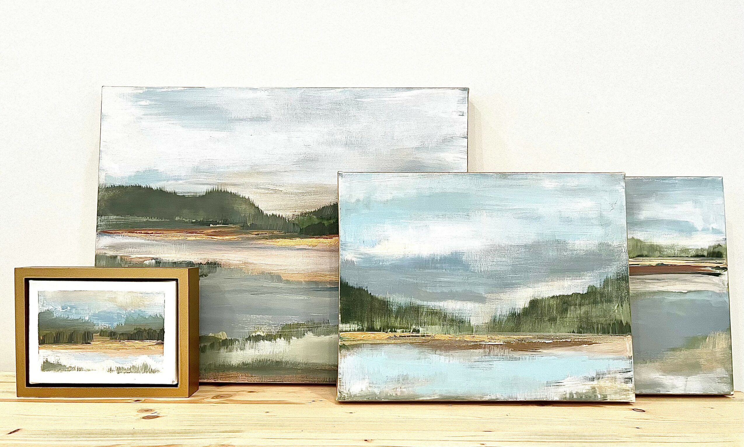 Four landscape paintings of lakes and mountains, with one framed, displayed on a light-colored surface against a white wall.