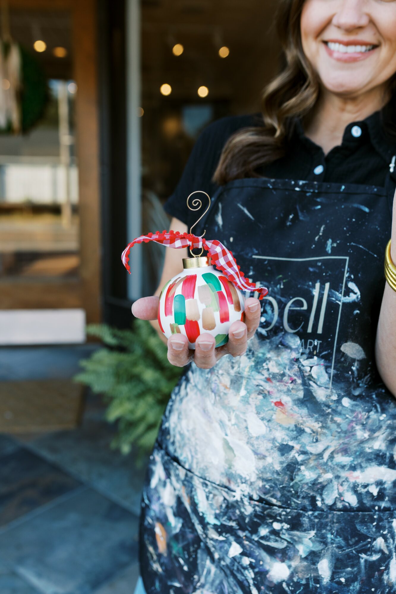 Person holding a decorated Christmas ornament with a red and white ribbon, smiling, wearing a black apron with paint splatters.