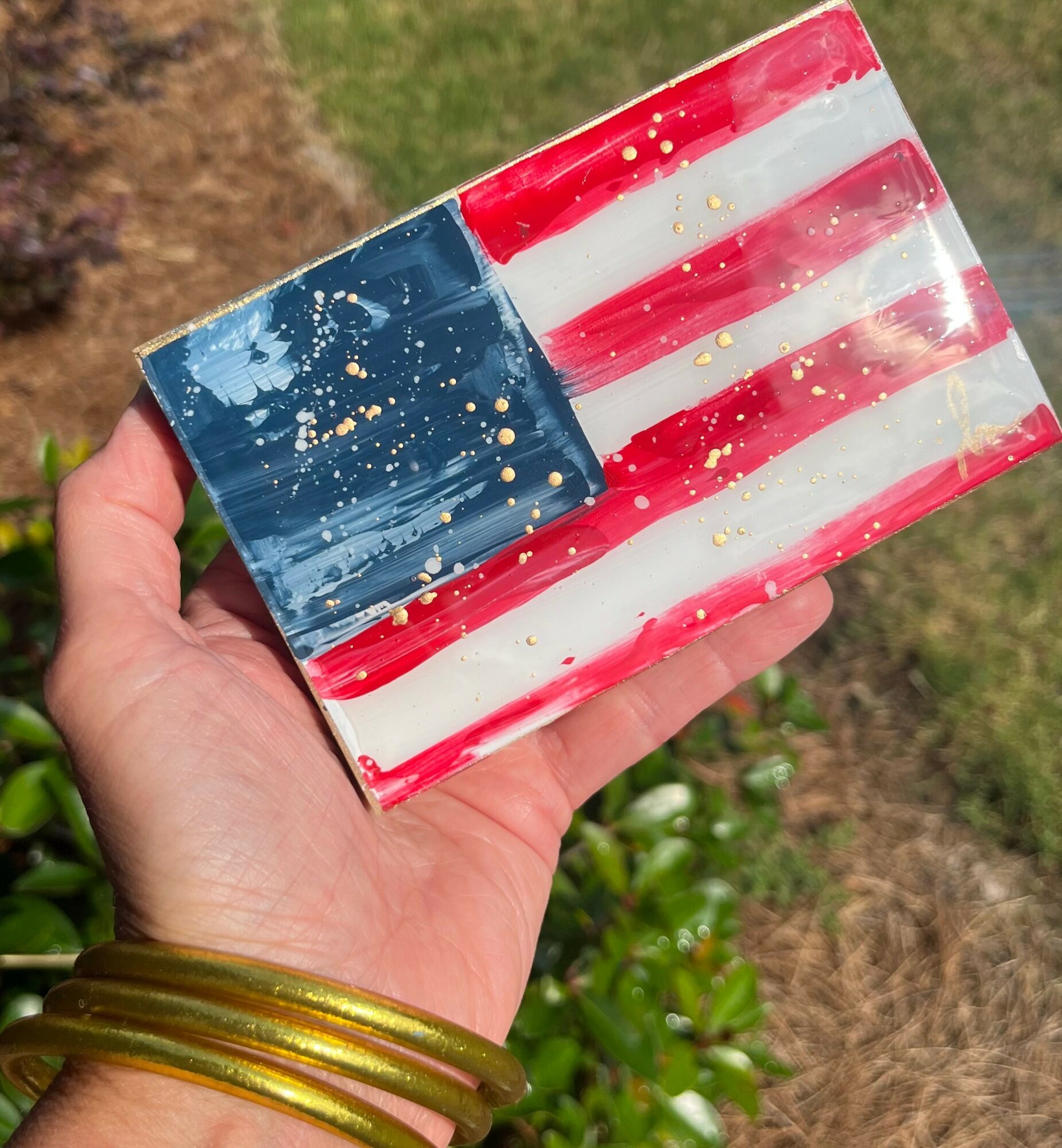 Hand holding a rectangular object painted with red, white, and blue stripes with gold splatters, outdoors with grass and plants.