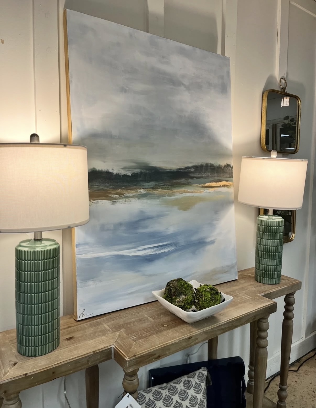 Large abstract landscape painting with sky, water, and land, flanked by two green ceramic table lamps on a wooden console table, with a bowl of moss in front.