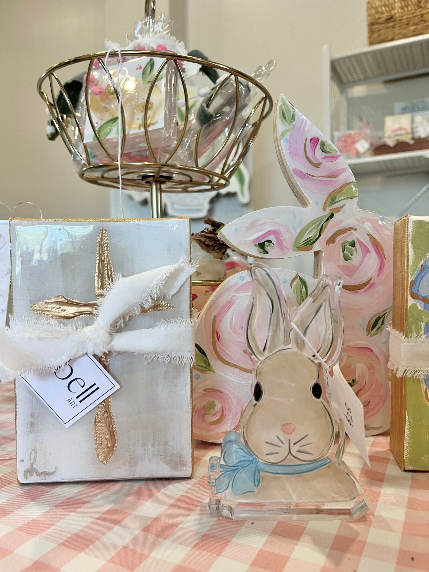 Decorative bunny figurine with a blue bow, gift box with a white bird, and a wire basket with flowers on a checkered surface.