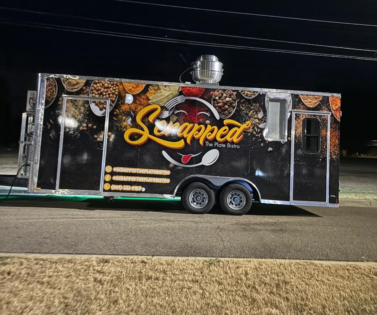 Food truck with a black background and colorful logo, parked on a street at night.