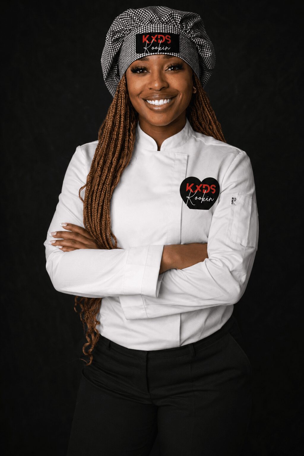 Woman in chef uniform with a hat, smiling, arms crossed, black background.