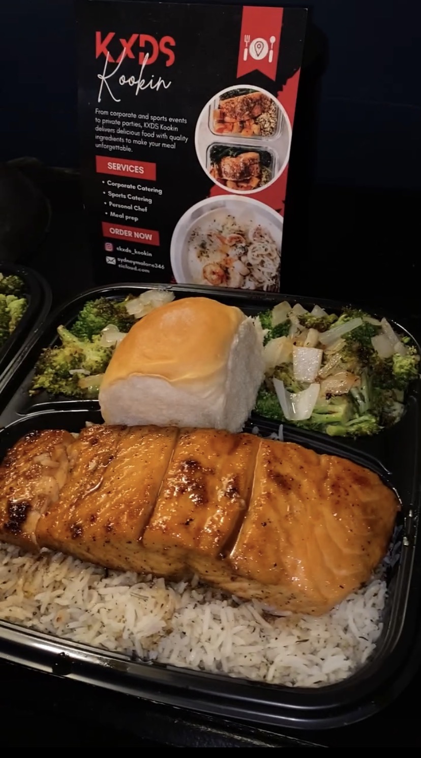 Meal with grilled salmon, rice, salad, and a dessert in a black container, with a menu in the background.