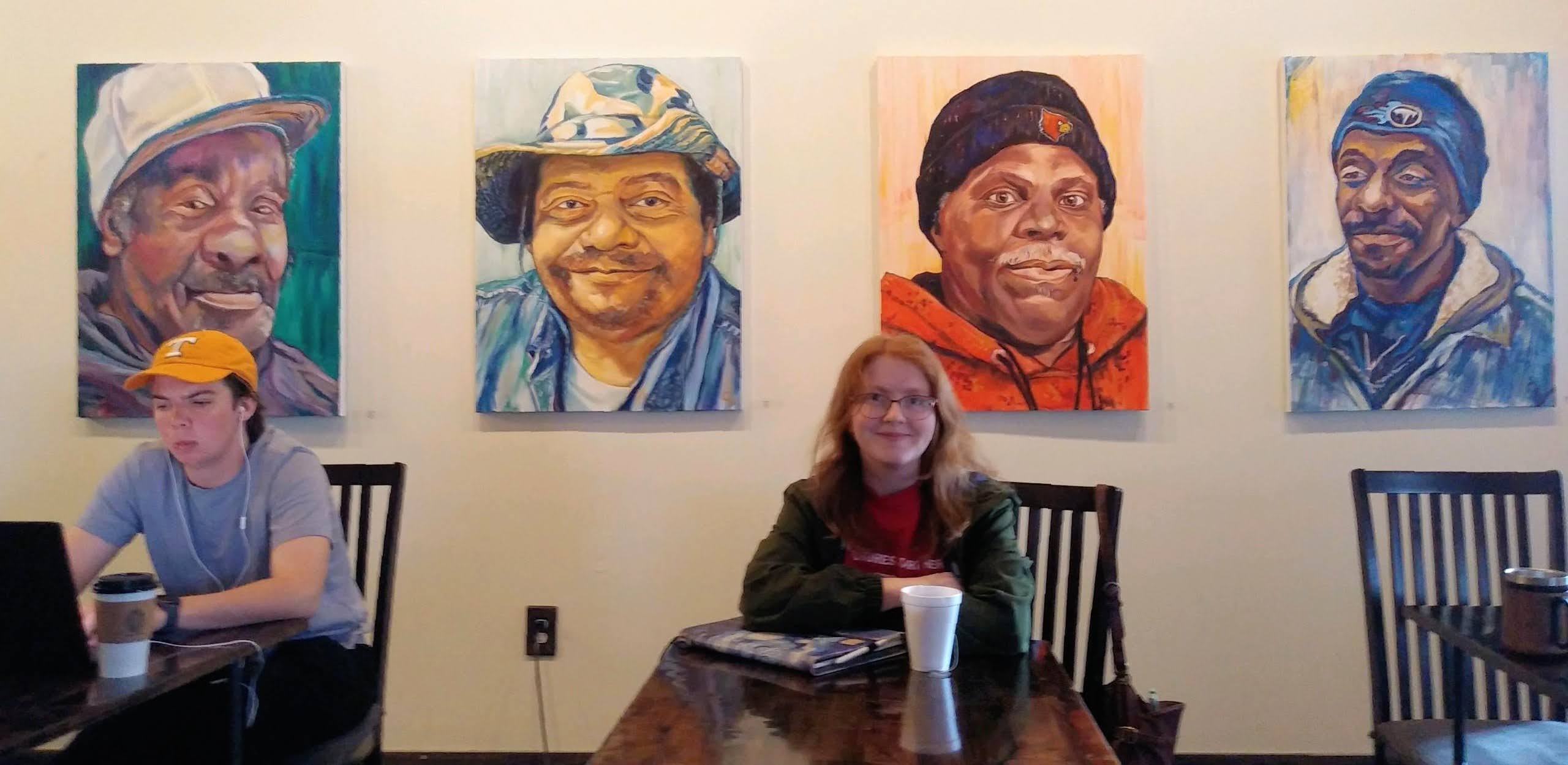 Four portraits of different people hanging on a wall, with three women sitting at a table in front.