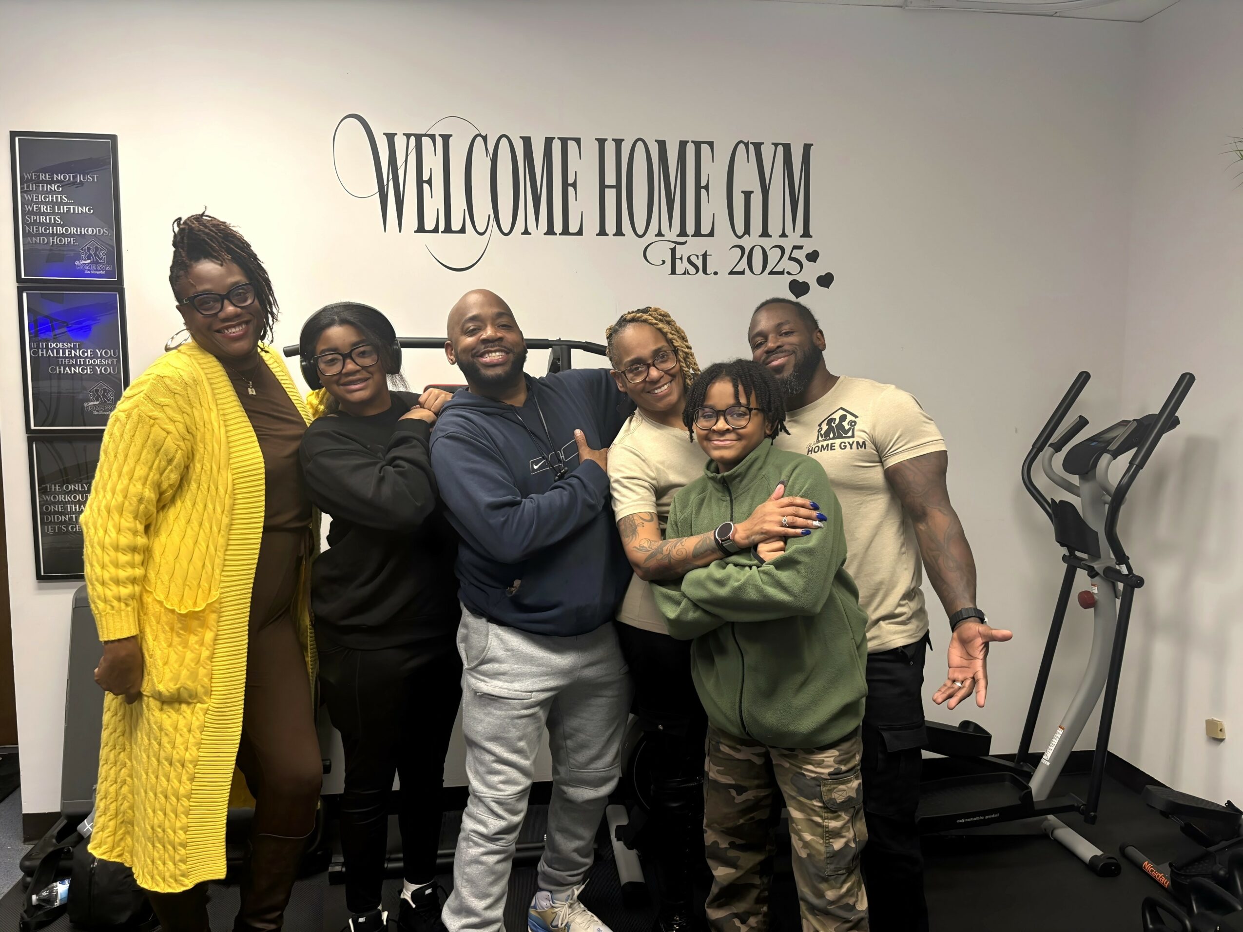 Group of six people smiling and posing in a gym, with a sign reading 'Welcome Home Gym Est. 2025' on the wall.