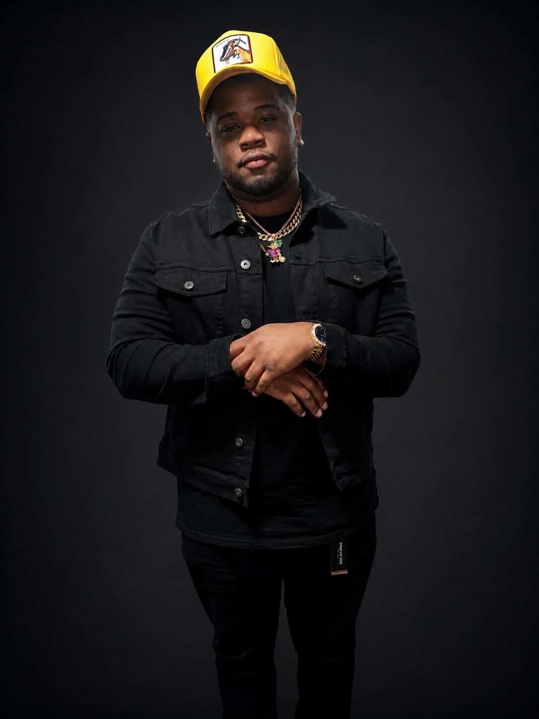 Person wearing a yellow cap, black jacket, and jewelry, standing against a dark background.