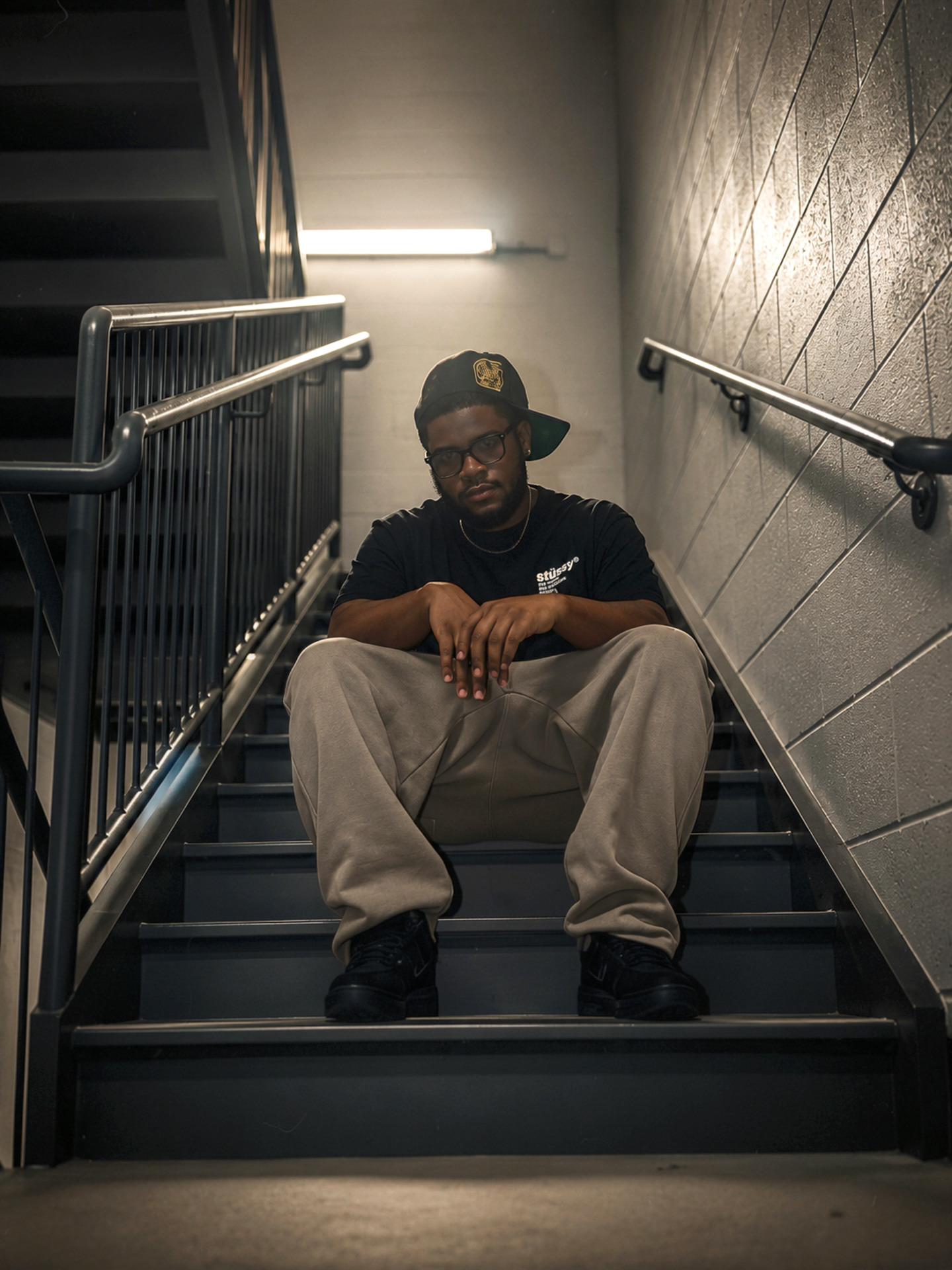 Person sitting on stairs with arms resting on knees, wearing a cap, glasses, black shirt, and beige pants, in an indoor setting.