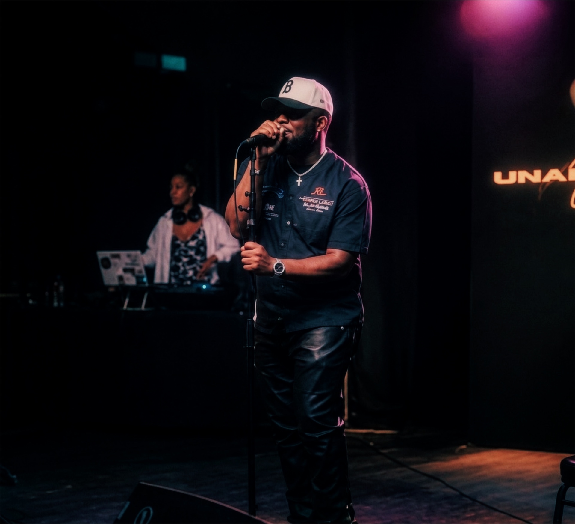 Man with a beard wearing a cap and dark clothing, holding a microphone on stage with DJ equipment in background.