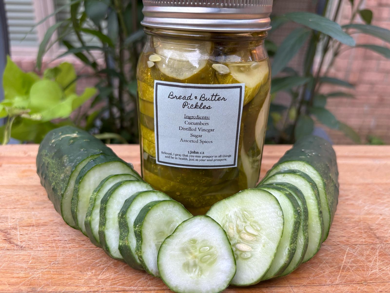 Sliced cucumber arranged in a semi-circle in front of a jar of pickles on a wooden surface, with green plants in the background.