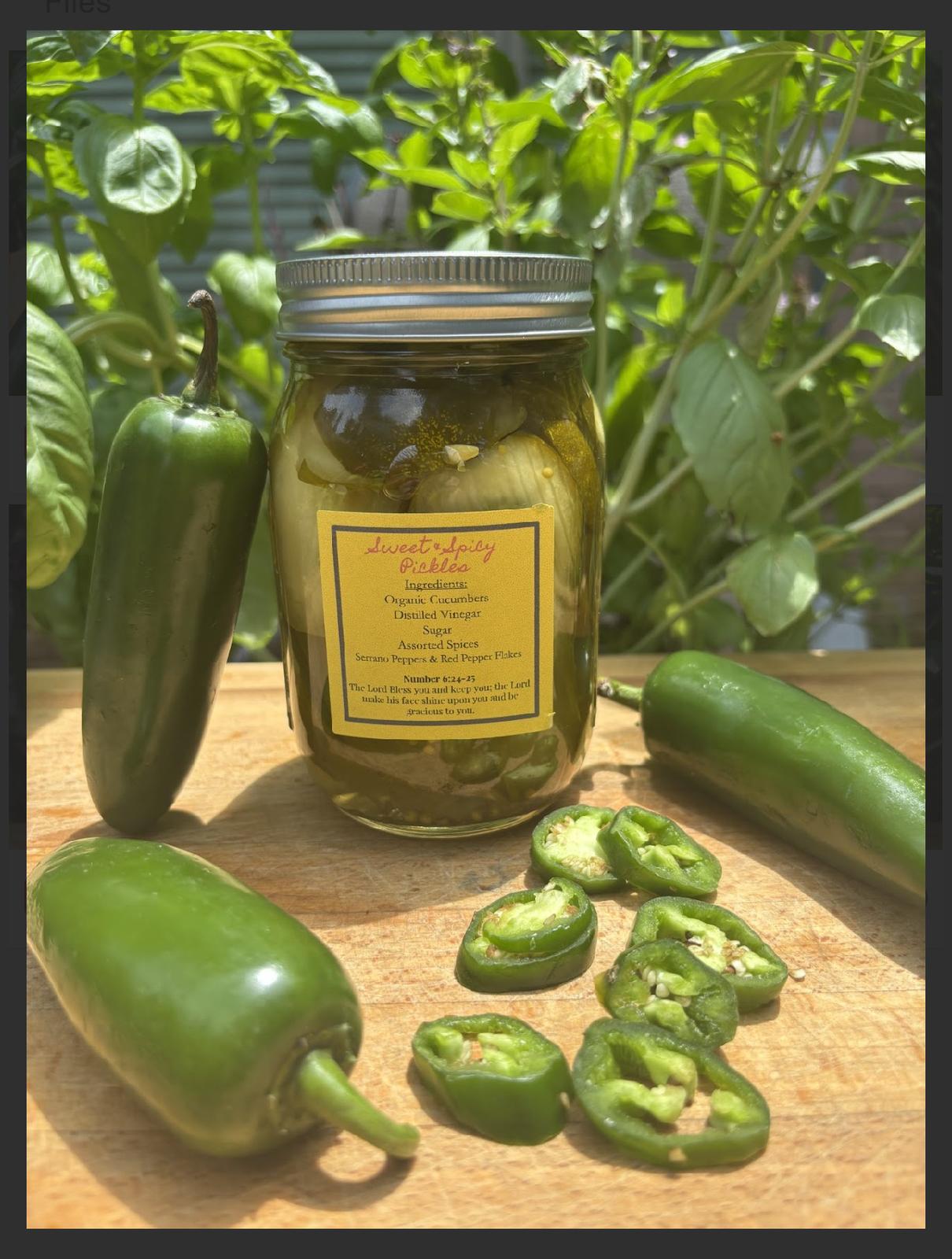Jar of pickles surrounded by fresh green peppers and sliced peppers on a wooden surface with green plants in background.