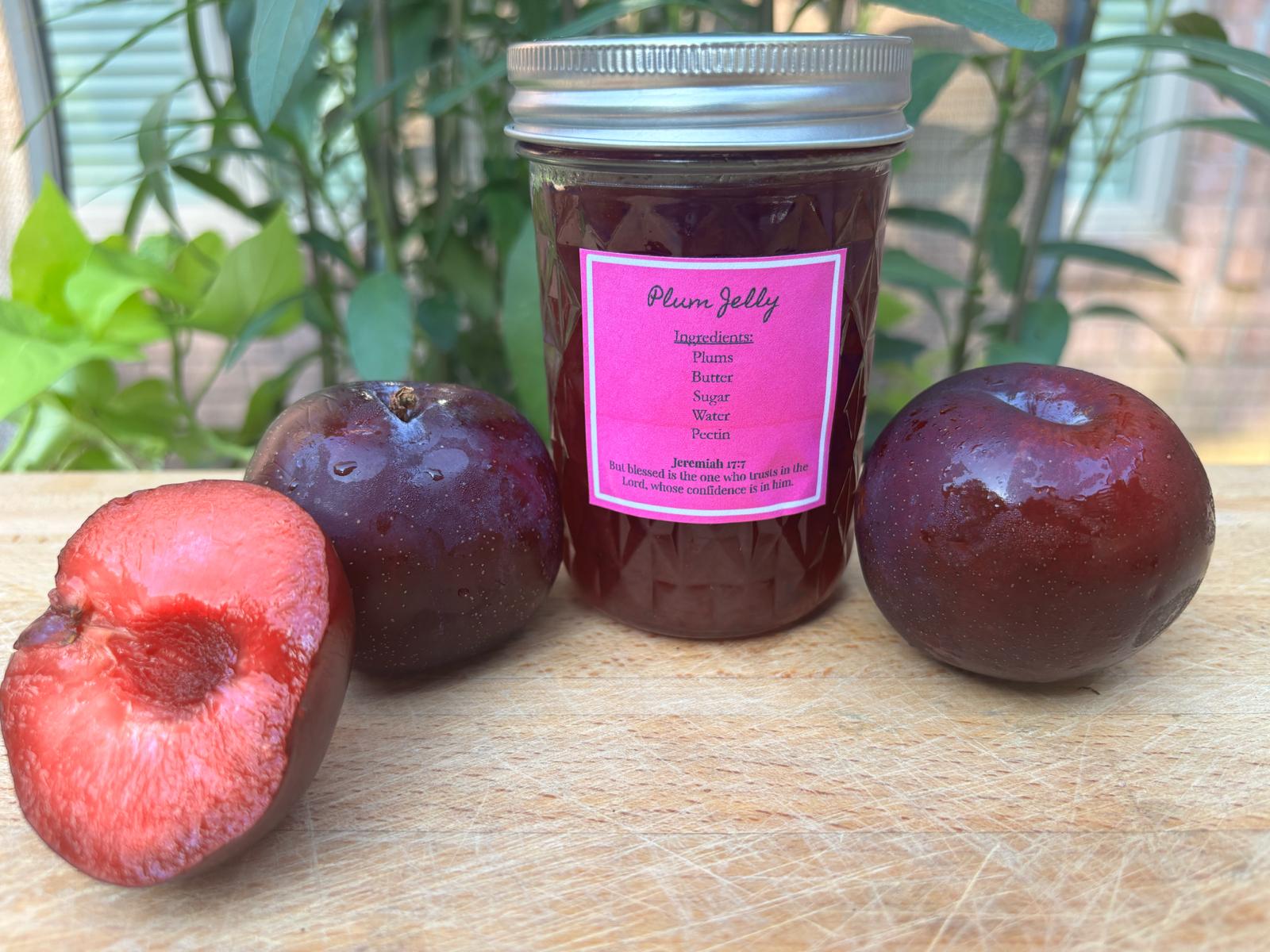 Three plums and a jar of plum jelly on a wooden surface with green plants in the background.