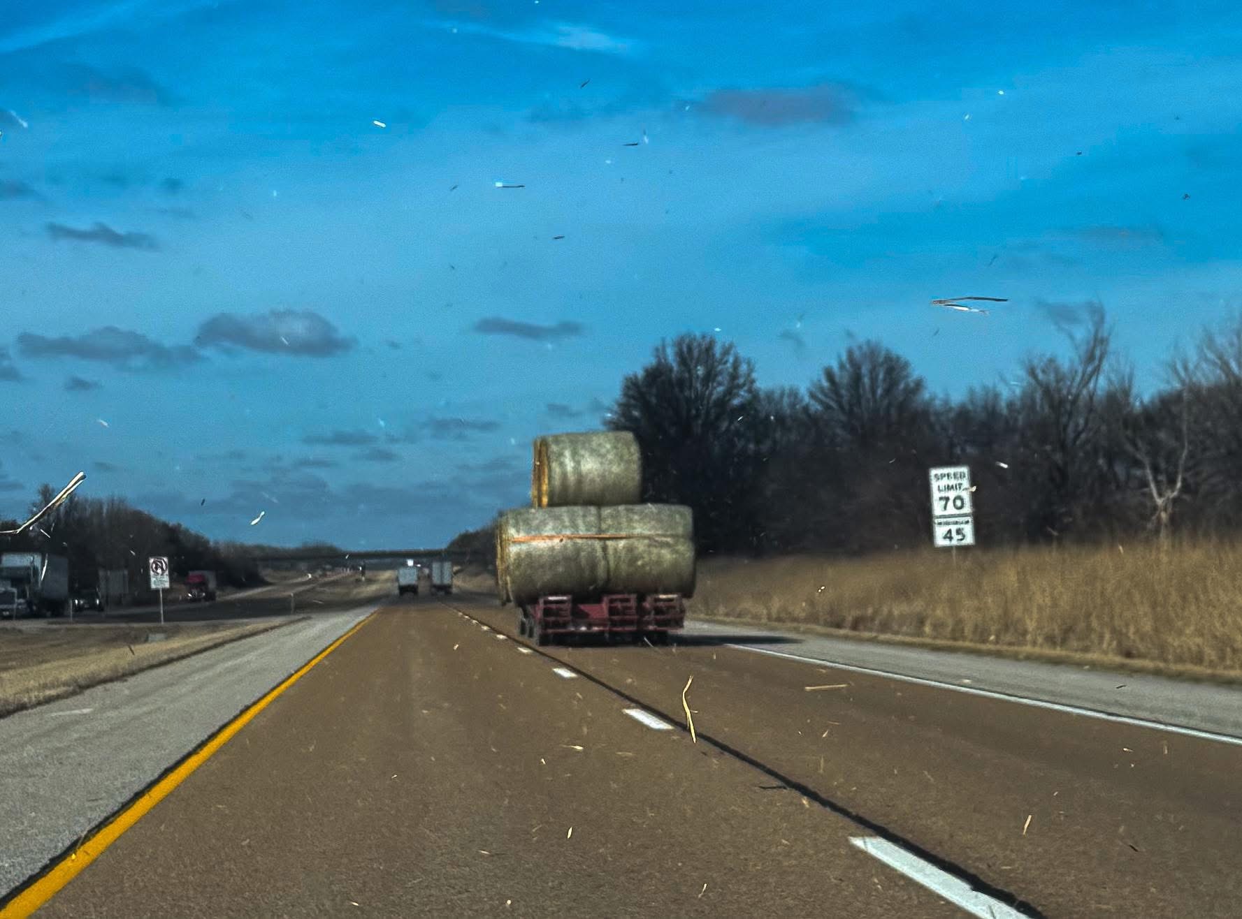 A truck on a highway carries large hay bales, with trees and a speed limit sign in the background.