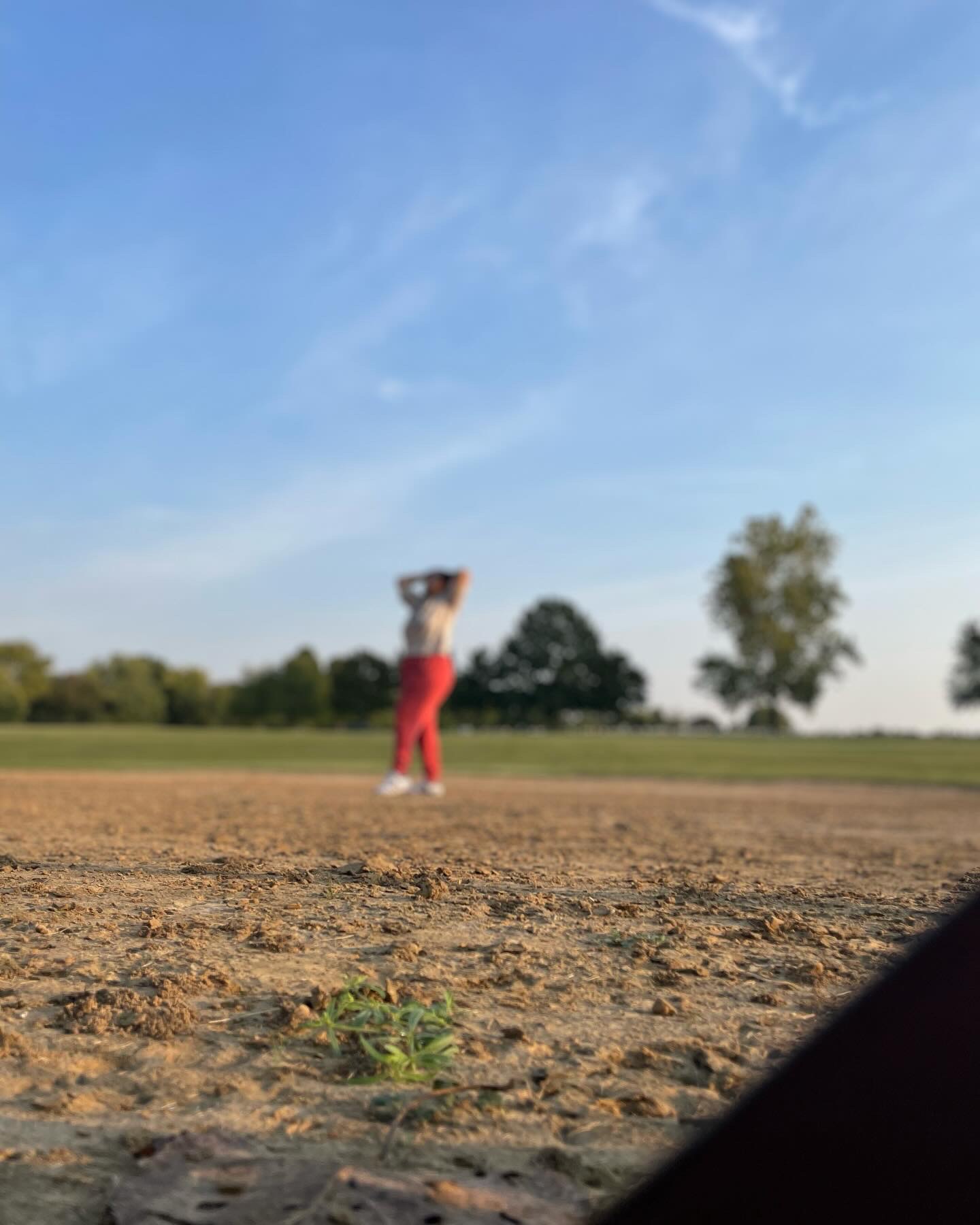Person standing on a dirt field with trees and blue sky in the background, holding an object near their head.