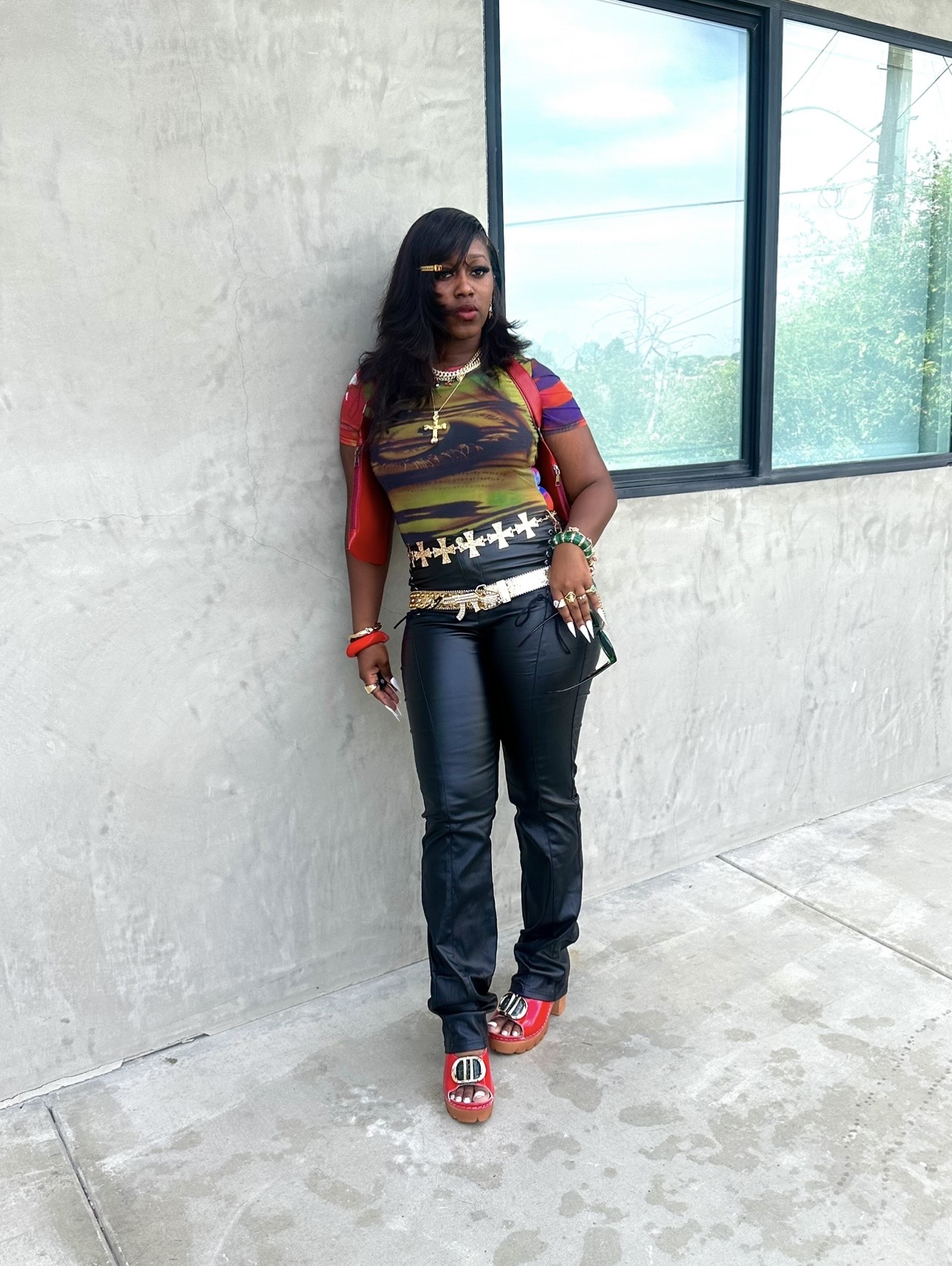 Woman standing indoors near a concrete wall and large window, wearing colorful top, jeans, and red shoes.