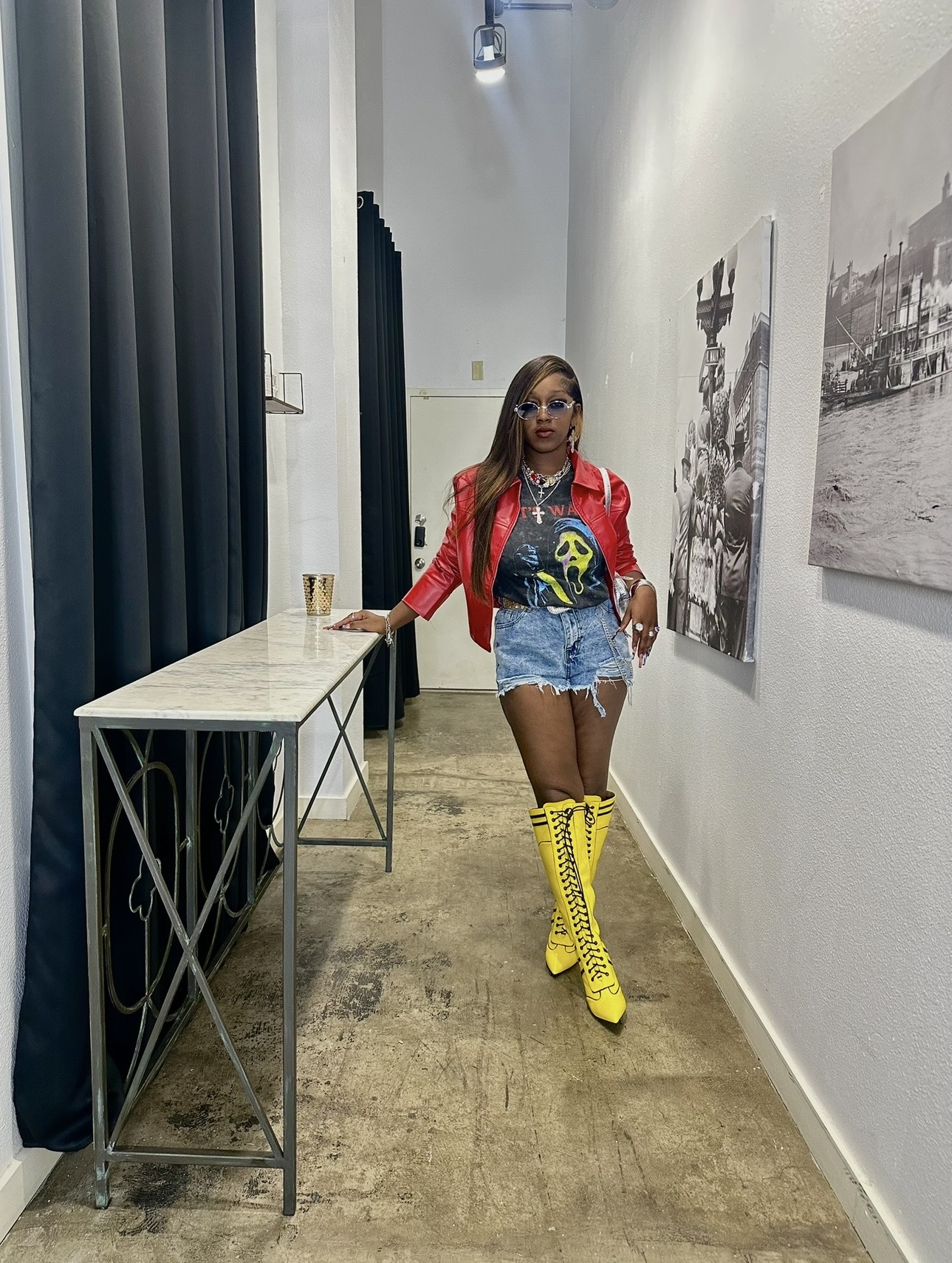 Woman in red jacket, sunglasses, denim shorts, and yellow boots standing in art gallery with black and white photos on wall.