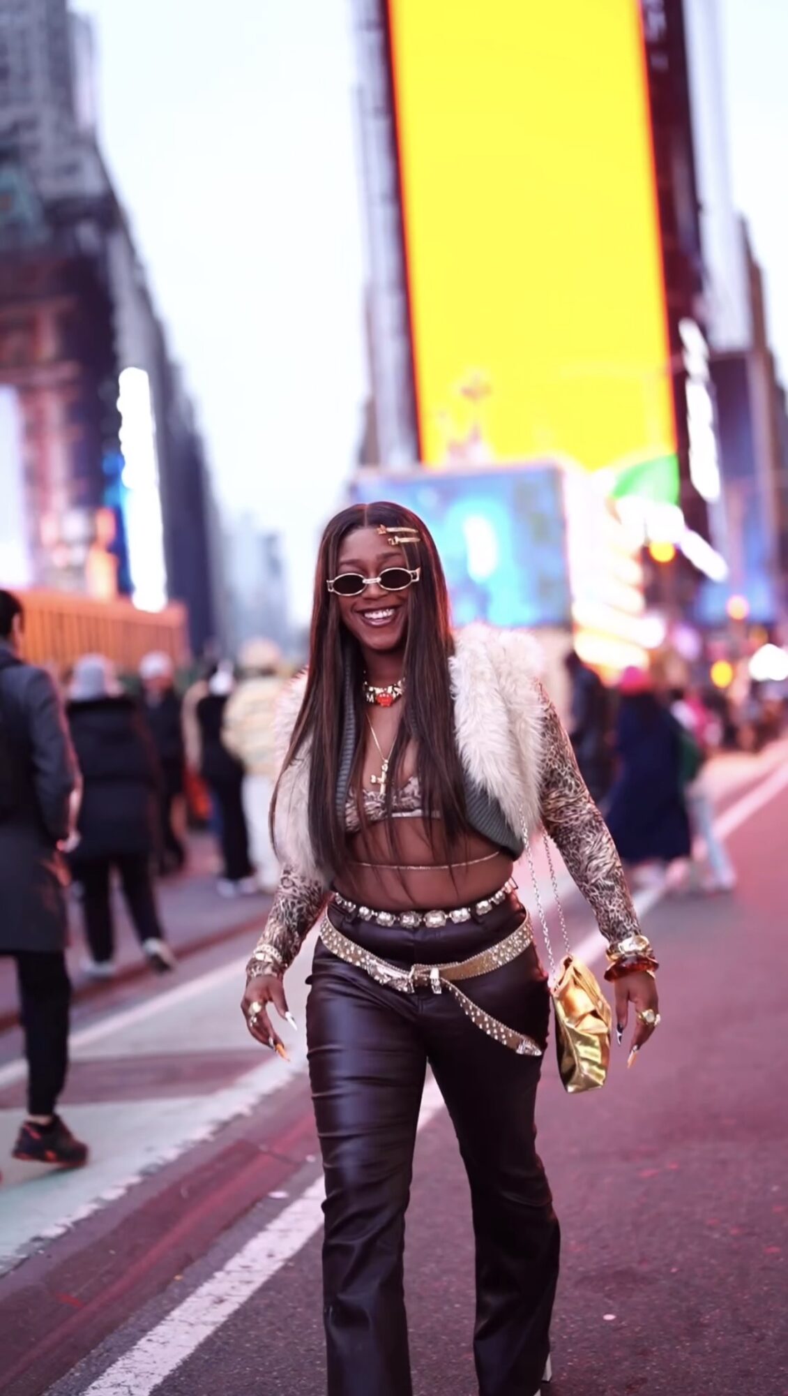 Woman smiling in Times Square with bright billboards, wearing sunglasses, a sheer top, and a fur jacket, holding a gold clutch.
