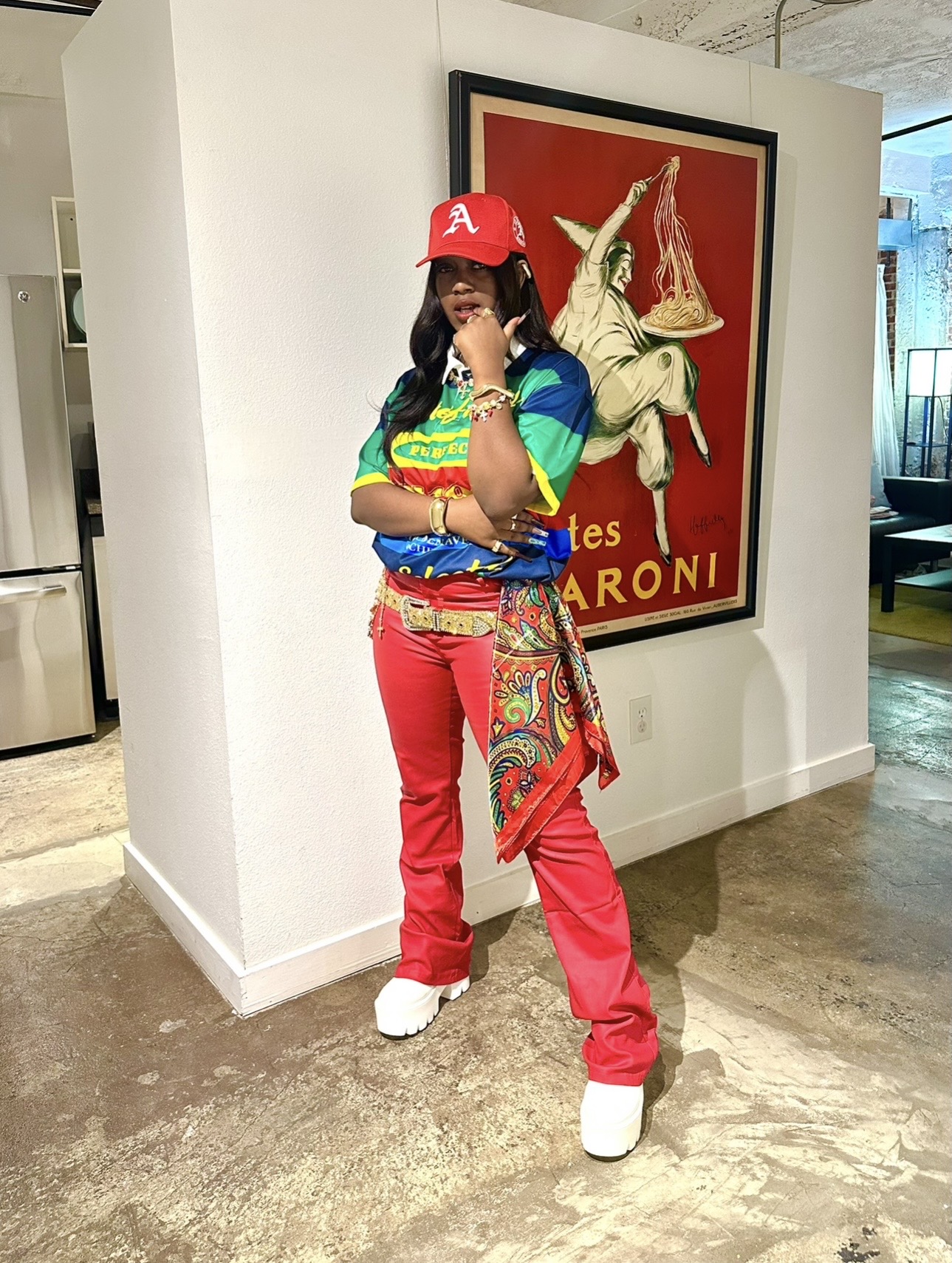 Person dressed in colorful clothing, red pants, and a red cap, standing in front of a framed poster of a horse.