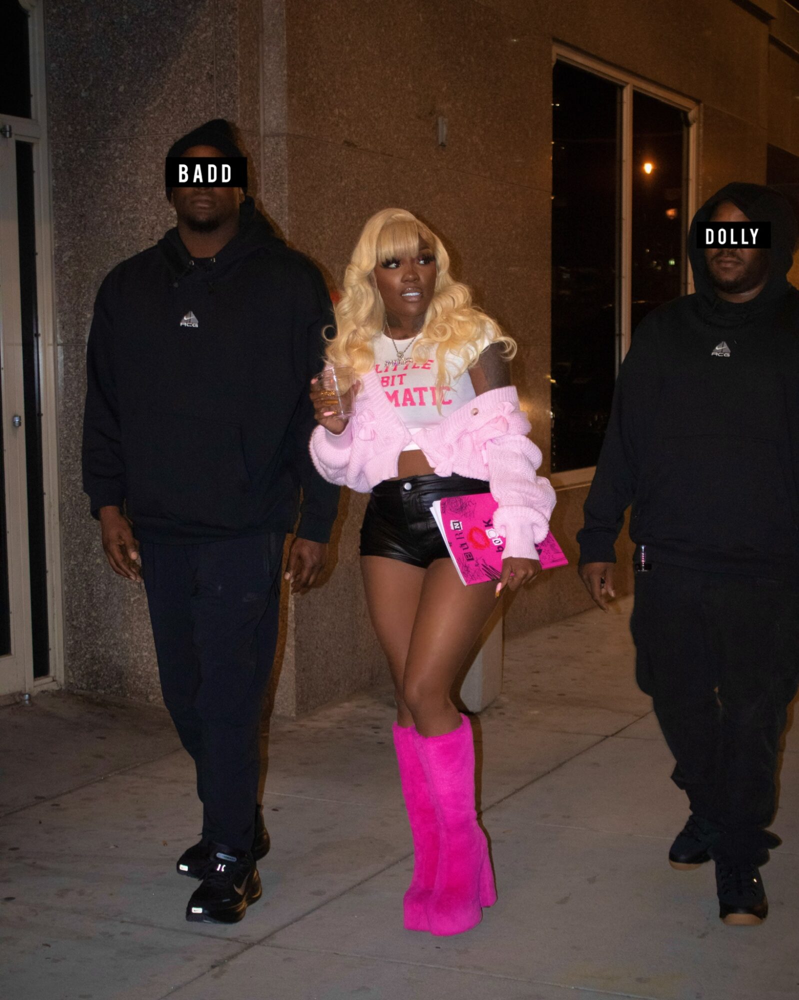 Person with long blonde hair wearing pink boots, shorts, and a pink jacket, walking between two men at night.