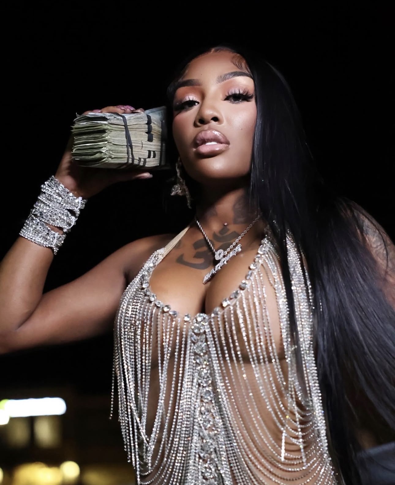 Woman with long black hair wearing jewelry, holding a stack of cash to her ear, against a dark background.