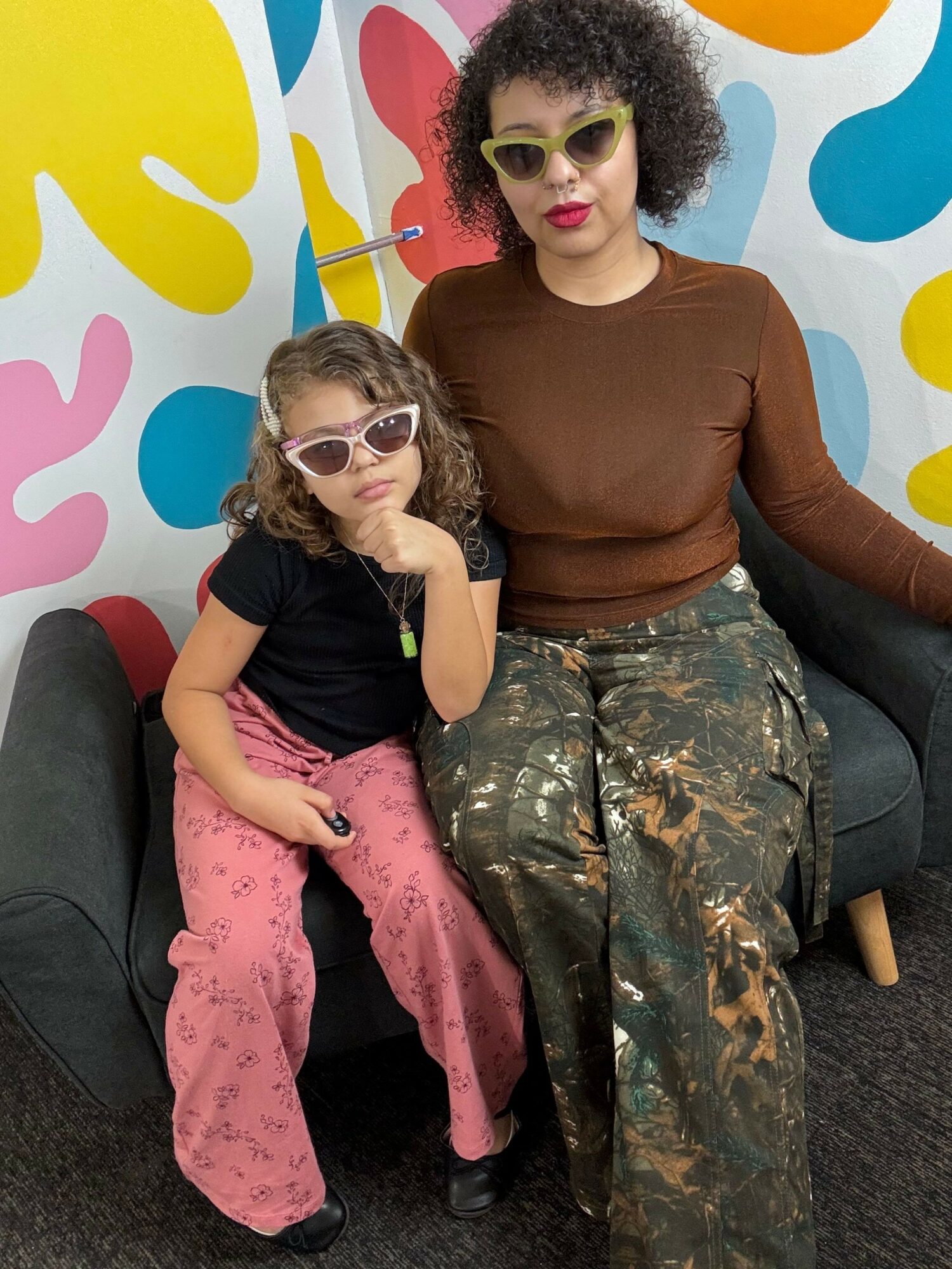 Two children with sunglasses sitting on a black couch, one with curly hair and the other with straight hair, in front of a colorful polka dot background.