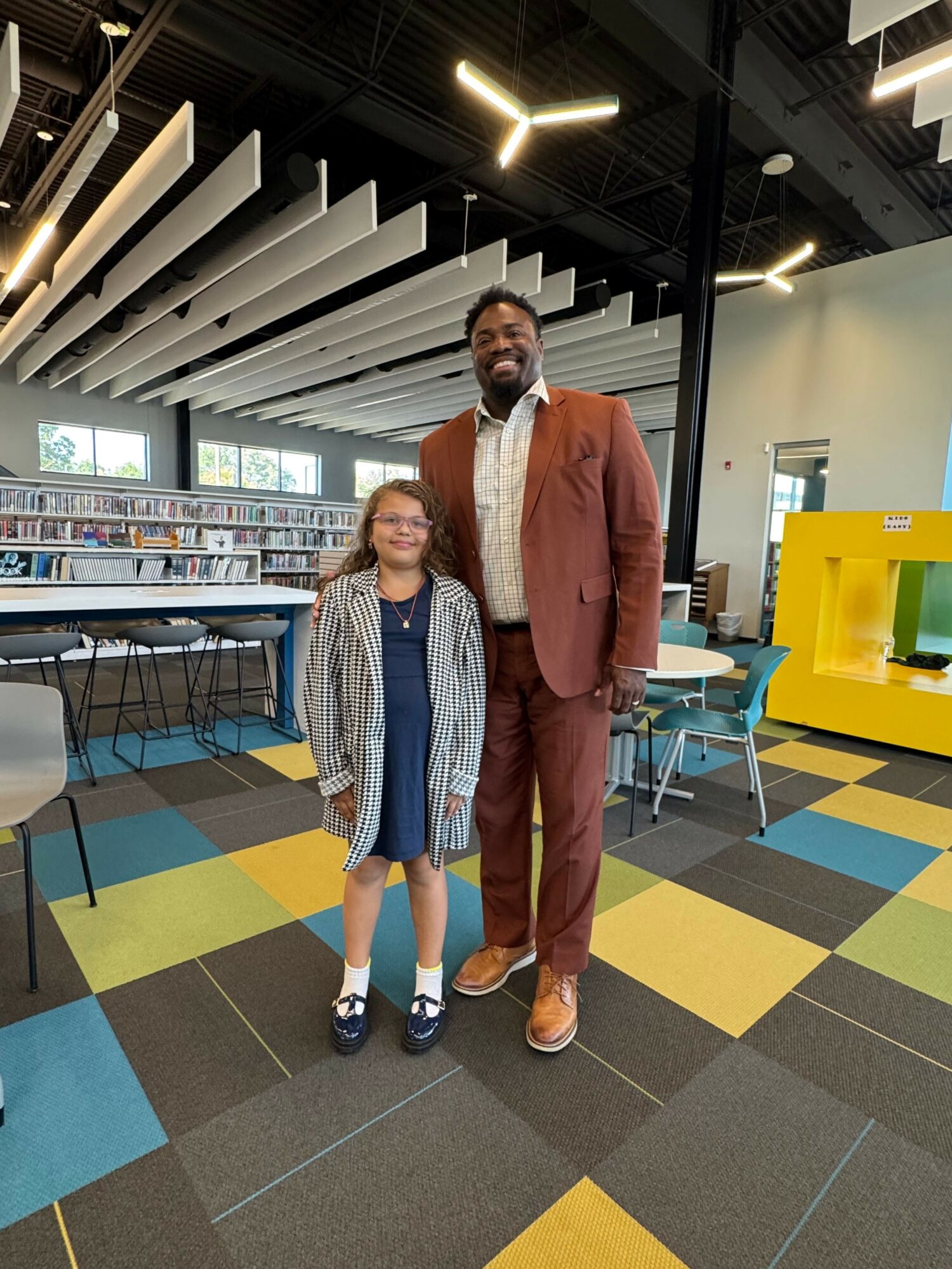 Two people standing in a modern indoor space with colorful flooring and large windows, one in a brown suit and the other in casual clothing.