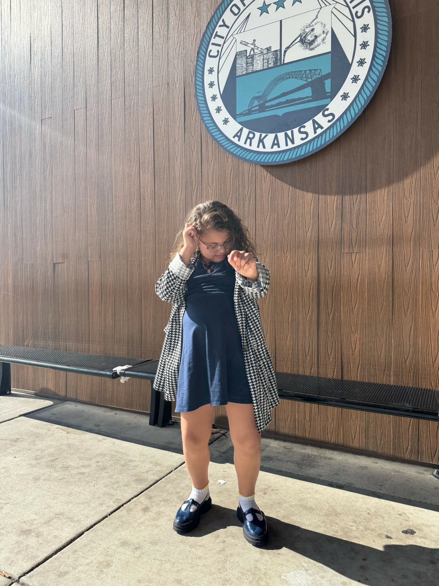 Girl standing in front of a wooden wall with a seal emblem, holding a phone to her ear, wearing a coat, shorts, and sneakers.