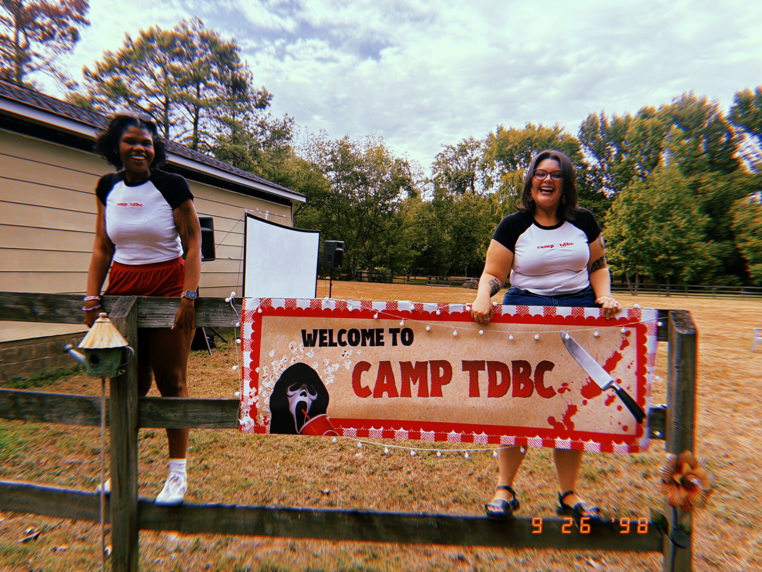 Two smiling women standing behind a camp sign at Camp TDBC, with trees and a building in the background.